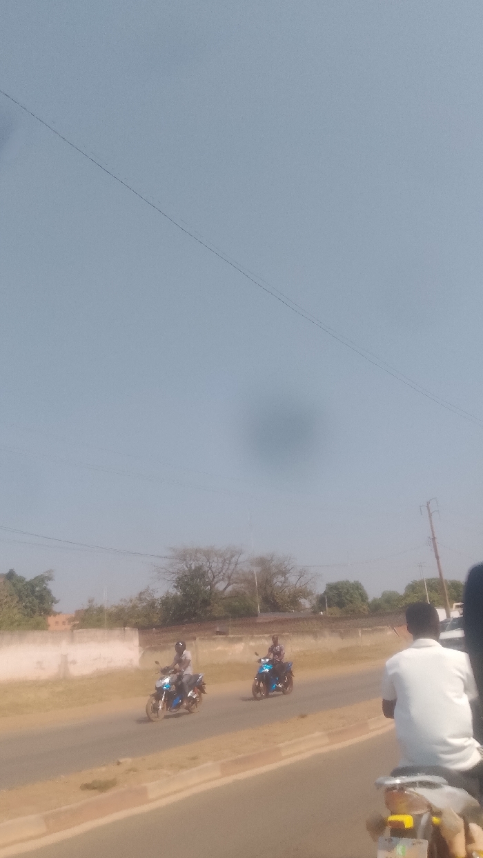 The image depicts a daytime street scene in Korhogo, Côte d'Ivoire, under a pale blue, hazy sky. The foreground shows the back of a person wearing a white polo shirt, seated on a scooter or motorcycle with yellow and black accents on the rear. This individual is positioned on the right side of the frame, facing away from the viewer. In the mid-ground, two individuals are operating motorcycles in the opposite direction. The motorcycle on the left is primarily blue and black, driven by a person wearing a light-colored shirt and dark pants, with a dark helmet. The motorcycle on the right is also blue and black, driven by an individual wearing a patterned shirt, dark pants, and a dark helmet. The road surface is asphalt, with a light-colored curb or median visible on the right. In the background, a low, light-colored wall or fence runs parallel to the road, behind which deciduous trees with sparse foliage and some green bushes are visible. A utility pole with overhead wires is present on the right side of the background. Buildings are partially discernible behind the trees and wall. A dark, circular smudge, consistent with a camera sensor artifact, is prominent in the upper center of the sky.