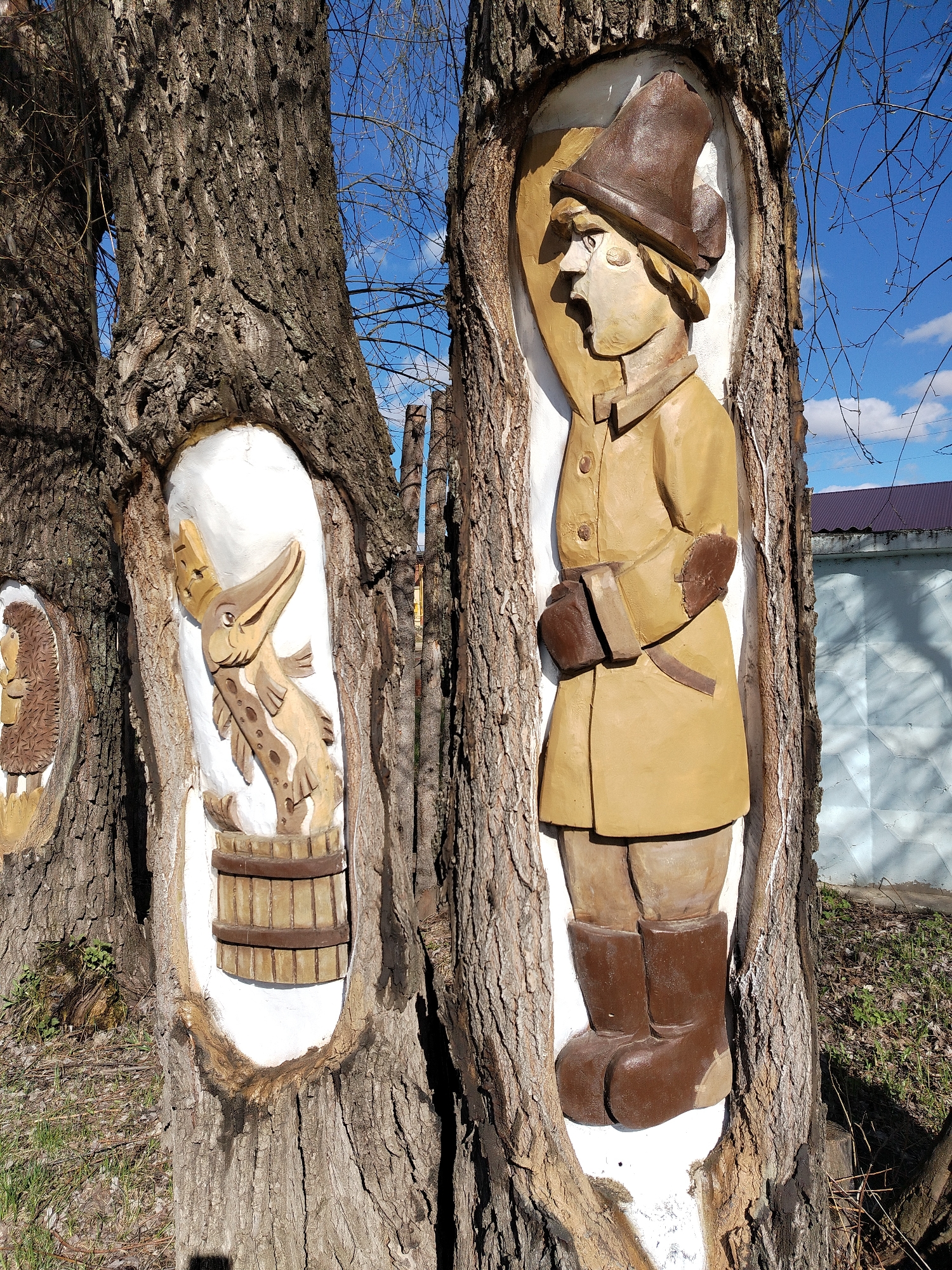 In the city of Chaikovsky, Russia, the image depicts a collection of wood carvings embedded into tree trunks. The carvings are painted in earth tones, with white paint surrounding each figure within the tree's hollow. The central figure, on the right, is a human figure wearing a hat, coat, gloves, and boots. To the left of the man is a carving of a creature, perhaps an alligator or crocodile-like figure holding a fish in its mouth, standing in a barrel. Further to the left, partially visible, is a carving of a hedgehog. The trees are thick and mature. The background shows glimpses of a blue sky with light clouds, a partial view of a building with a purple roof and a light blue wall, and bare tree branches. Ground foliage is present at the base of the trees.