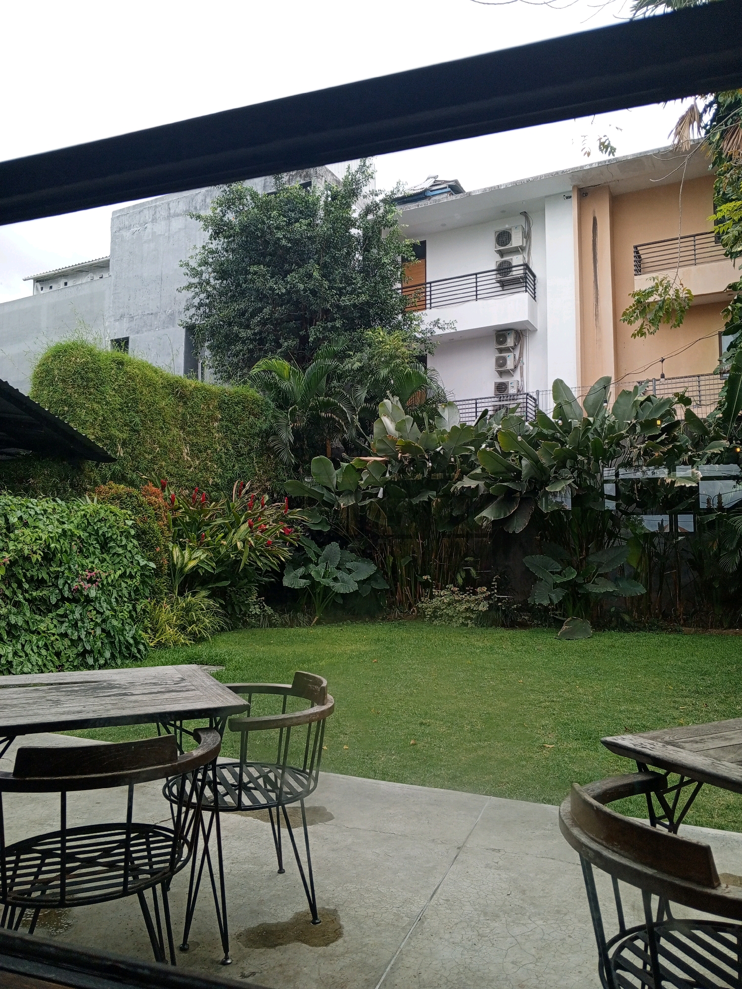 The image depicts an outdoor seating area at a cafe or restaurant in Denpasar, Indonesia, with a lush garden and a multi-story building in the background. In the foreground, a weathered wooden table and two metal-framed chairs with wooden seats are positioned on a concrete patio. The patio shows signs of recent rain, with puddles of water present.
Behind the patio, a vibrant green lawn stretches out, leading to a dense, tropical garden filled with a variety of plants. Notable vegetation includes tall, leafy plants with large leaves, bamboo, and flowering plants with red blossoms. The garden appears well-maintained and lush.
In the background, a multi-story building is visible. The building has a modern design with white and peach-colored facades. Several air conditioning units are mounted on the exterior walls, and balconies with black metal railings are present. The upper floors suggest residential or commercial spaces. The sky is overcast, indicating a cloudy or possibly rainy day. The overall scene conveys a serene and tropical ambiance.