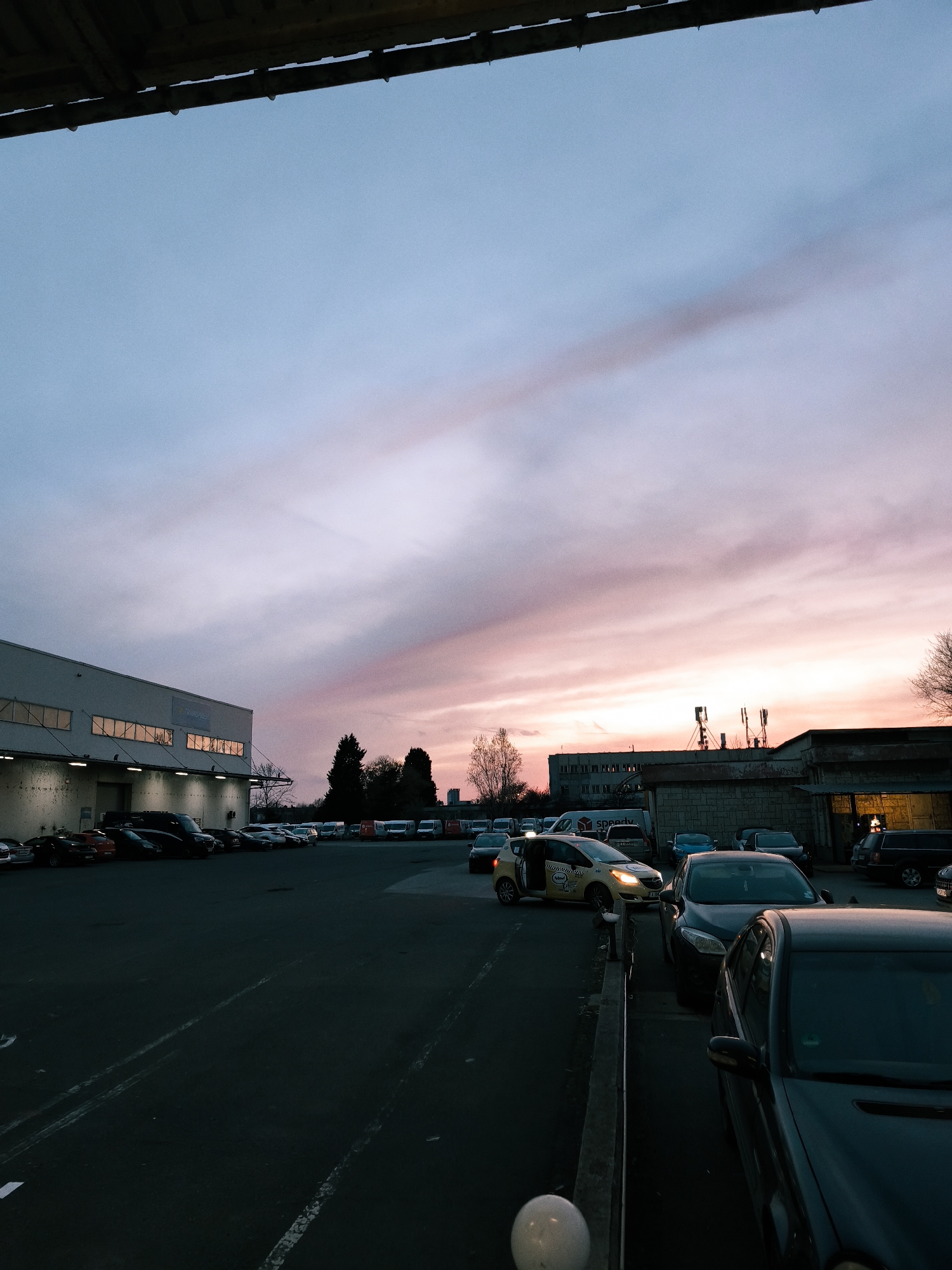 The image captures an outdoor parking lot or industrial area in Burgas, Bulgaria, during what appears to be twilight or sunset. The sky is a prominent feature, transitioning from a light blue at the top to vibrant pink and purple hues closer to the horizon, with subtle cloud formations stretching across. A dark, wooden or metallic beam, possibly part of an overhead structure, frames the very top of the image.
On the left, a large, rectangular, light-colored industrial building dominates the mid-ground. Its upper section features several rectangular windows reflecting the soft light, while an overhang below is illuminated by multiple downward-facing lights, casting a glow on the wall and parked vehicles. Further right, a collection of smaller, older-looking buildings with various textures, including one with exposed brickwork, stand against the colorful sky. Antennas or communication towers are visible on the roof of one of these structures. Bare trees with intricate branch patterns are scattered throughout the scene.
The parking lot itself is filled with numerous vehicles. In the foreground, several dark cars are parked, with a white spherical light fixture partially visible at the bottom left. Towards the center-left, a yellow taxi with "Taxi" written on its side, and partial text "Schwalbe," is prominent with its headlights on, appearing to be in motion or recently stopped. Behind it, a dark van and other cars are parked. In the distant background, rows of white and red vans, including one with the "SPEEDY" logo (a Bulgarian courier company), suggest a commercial or logistics hub. The asphalt ground of the parking lot shows faint white markings. No people are explicitly visible, contributing to a serene and transitional atmosphere at the day's end.