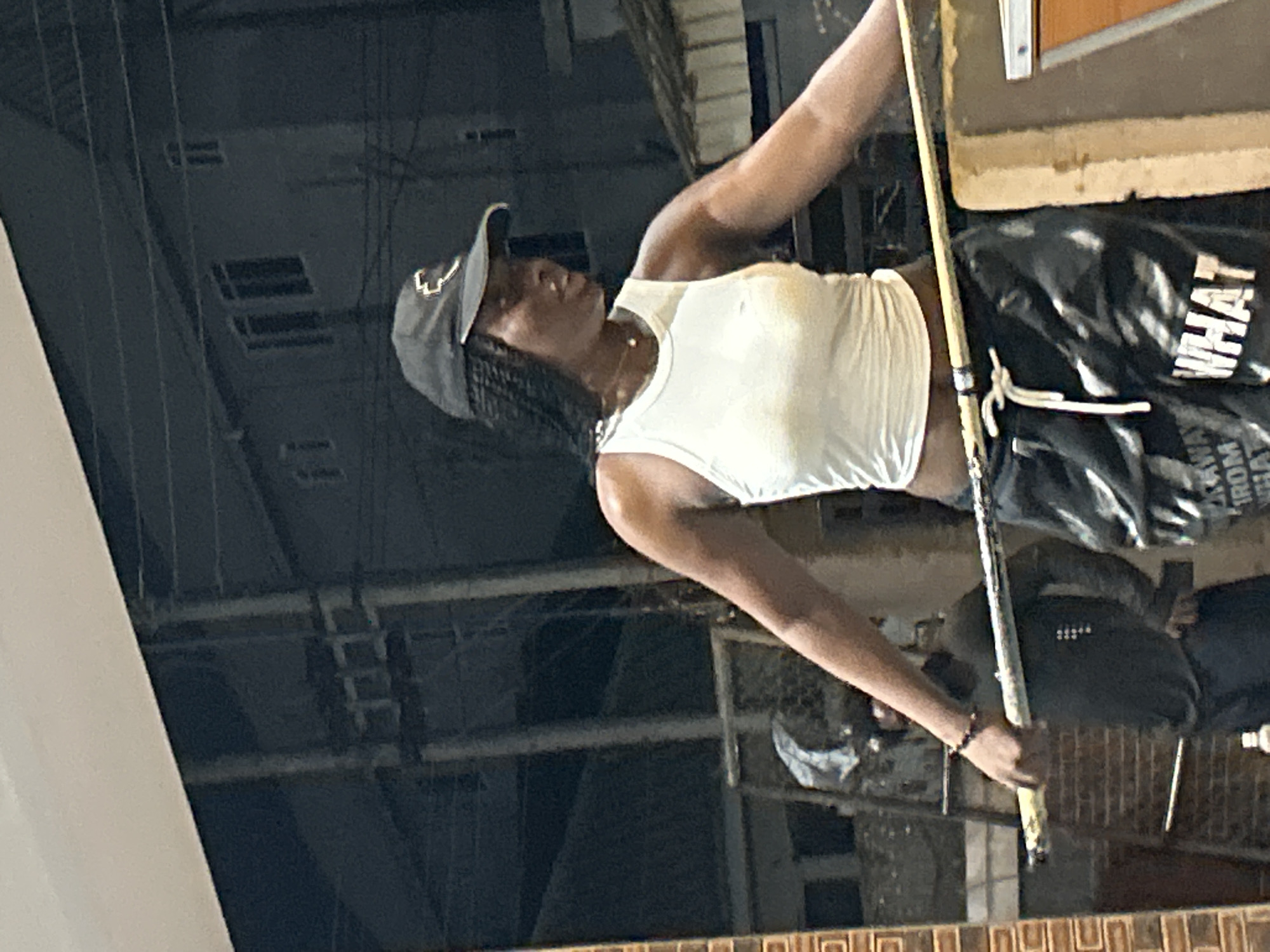 A woman wearing a white tank top and black baseball cap stands outdoors, holding a pool cue. She has braids and a thin necklace. She is wearing black and white patterned pants with the word "WHAT" visible on the right leg. Behind her, a man in a dark shirt is visible, and in the background, there are buildings and electrical wires, suggesting an urban or semi-urban environment. The lighting suggests it is either evening or nighttime.
