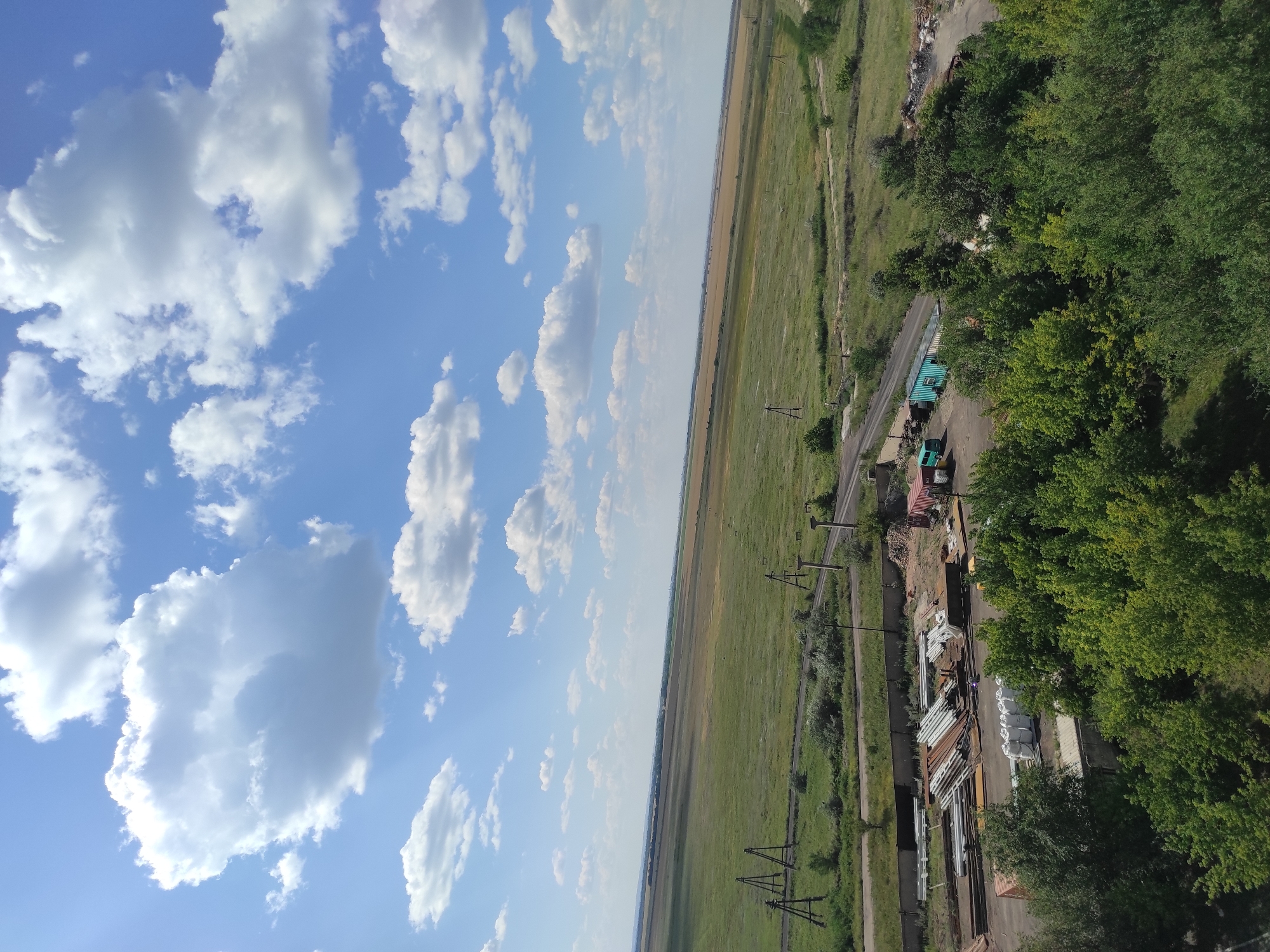 The image shows a wide, expansive landscape under a bright blue sky with scattered white cumulus clouds. The foreground is dominated by lush green trees. Beyond the trees, there is an industrial or storage area featuring stacks of metal pipes, construction materials, white sacks, and shipping containers. A road or path winds through this area. Further into the distance, the landscape transitions into vast agricultural fields, appearing green and yellow, with hints of brown earth. The horizon line is low, emphasizing the vastness of the sky and fields. There are no discernible people in the image. The overall impression is of a rural or semi-industrial area with a clear, sunny day. The location is stated to be the city of Kyzyl, Kazakhstan.