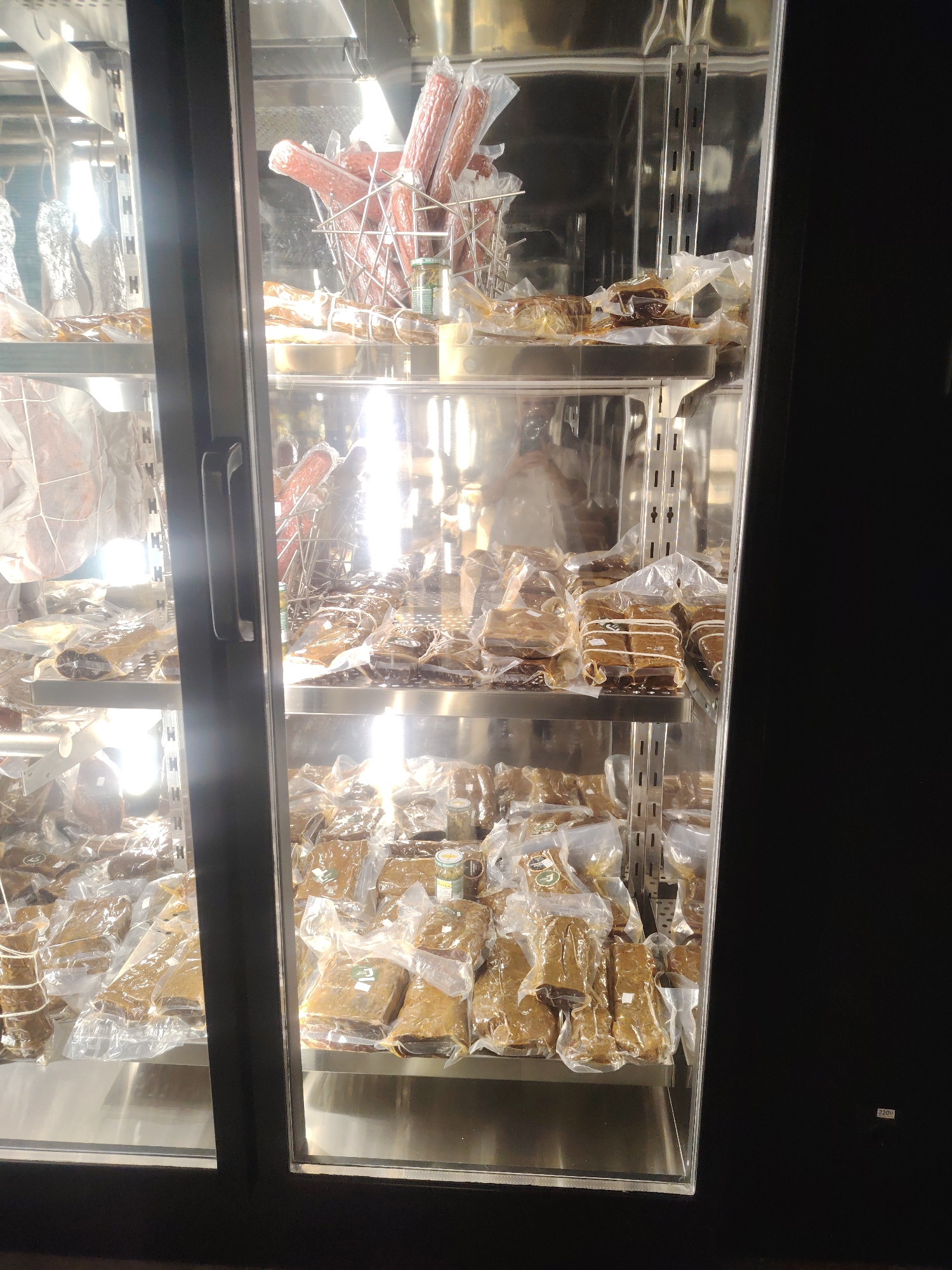In Konigil, Uzbekistan, a brightly lit display refrigerator contains various packaged food items, primarily cured meats and what appear to be preserved sausages. The refrigerator has glass doors and multiple shelves, all filled with products sealed in transparent plastic. On the top shelf, several long, cylindrical sausages are arranged in a metal basket. Below this, multiple rows of smaller, rectangular packages are stacked, some tied with string. The background of the refrigerator is reflective, likely stainless steel, showing distorted reflections of the food and the lighting. The overall impression is a well-stocked display of preserved food items.