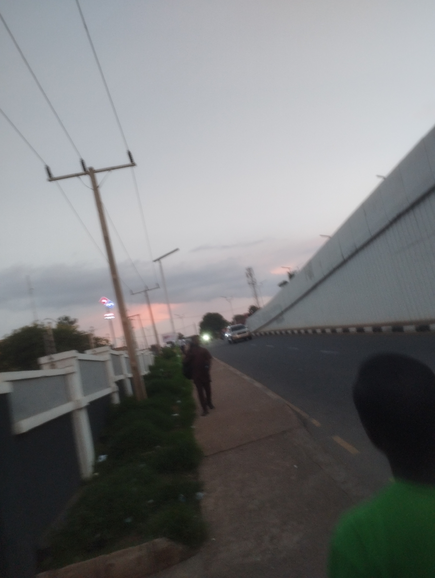 The image depicts an outdoor scene, likely at dusk, with a roadway and a pedestrian walkway. A large, elevated concrete barrier with black and white markings runs along the right side of the roadway. On the left side of the walkway, there is a low gray fence with white trim and some greenery. Several utility poles with wires are visible along the walkway, and one pole has a illuminated sign with red and blue lights.
Several individuals are present. One person, wearing dark clothing, is walking on the pedestrian walkway. In the foreground, on the right side of the image, the back of another person's head and shoulders are visible; they are wearing a bright green shirt. Two vehicles are on the roadway, one appears to be a silver sedan, and another is partially visible. The sky is overcast with hints of pink and orange from what appears to be a sunset. The overall impression is a scene on the outskirts of a city, possibly near an overpass or elevated section of road.
This scene is consistent with locations in Jos, Nigeria, particularly areas with modern infrastructure development. The presence of the illuminated sign could indicate a commercial area.