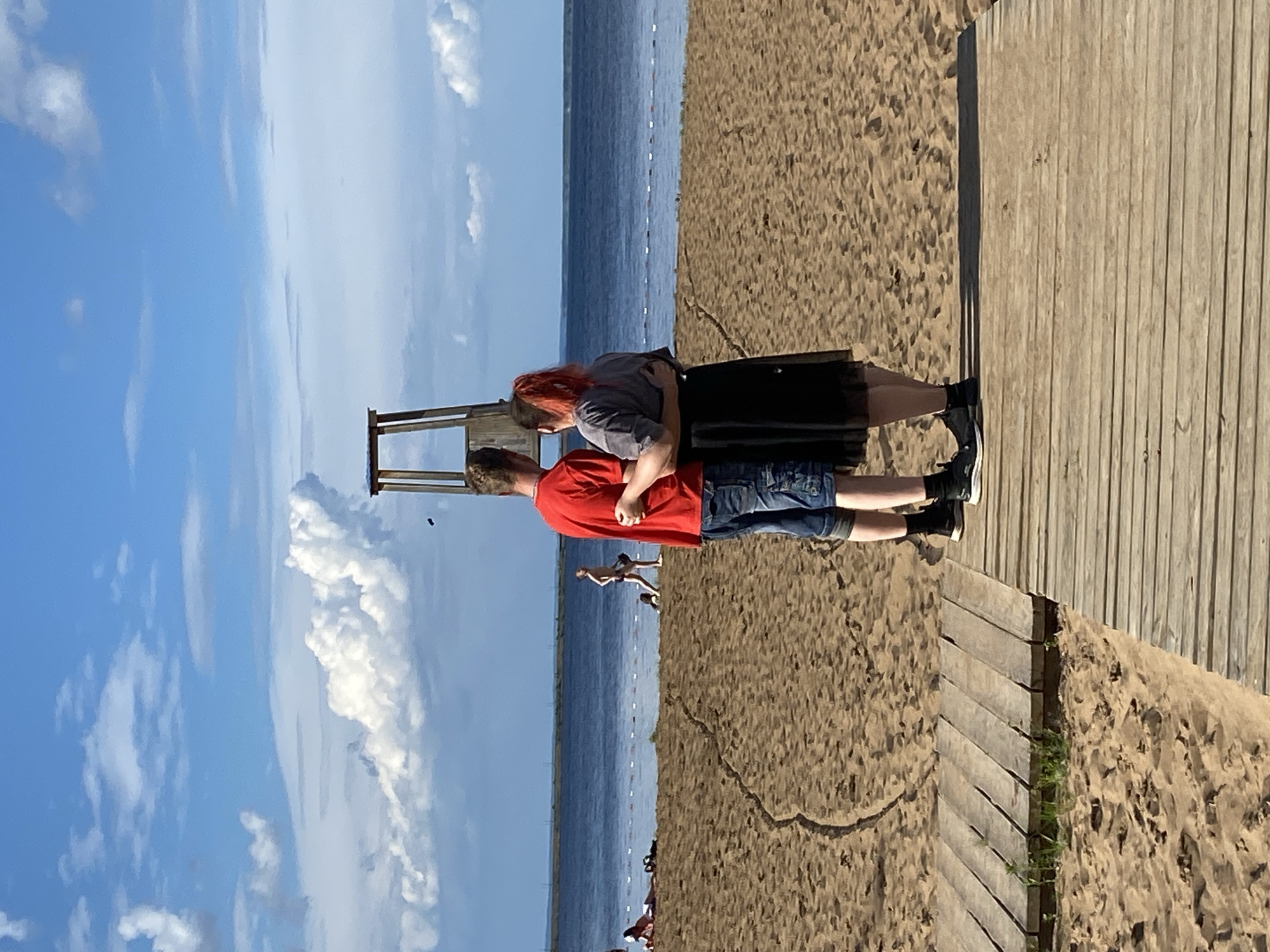 The image depicts a bright, sunny day at a beach, likely in Chaikovsky, Russia, given the context. Two individuals are the central focus, positioned on a wooden boardwalk that runs alongside a sandy beach. The person on the left, with striking red hair, wears a gray top and a black skirt, and is embracing the person on the right. The person on the right wears a red t-shirt and denim shorts. Their stance suggests an intimate moment, possibly a hug or a close conversation.
Beyond the boardwalk and the beach, a vast expanse of blue water stretches towards the horizon, where it meets a clear blue sky dotted with fluffy white clouds. The water is calm, with a thin white line of buoys marking a boundary in the distance. The sandy beach is textured, showing the imprint of footprints and natural contours.
Further in the background, several other beachgoers can be seen in the distance, some appearing to be walking or sunbathing. One individual stands out on the beach, closer to the water, and there are hints of other people lying down on the sand. The time of day appears to be late morning or early afternoon, judging by the strong sunlight casting distinct shadows. The weather is clearly pleasant and warm, ideal for a beach outing. The overall mood conveyed is one of relaxation and togetherness. No visible text is present in the image.