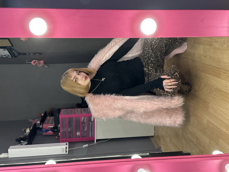 A person with blonde, blunt-cut bangs and a black turtleneck poses in a mirror's reflection. A light pink faux fur coat drapes over them, complementing their leopard print pants and matching booties. They are accessorized with a silver cross necklace. The backdrop reveals a pink makeup organizer, a white cabinet, and a grey wall. The mirror frame, painted a vibrant pink and lined with bright lights, adds a theatrical flair.