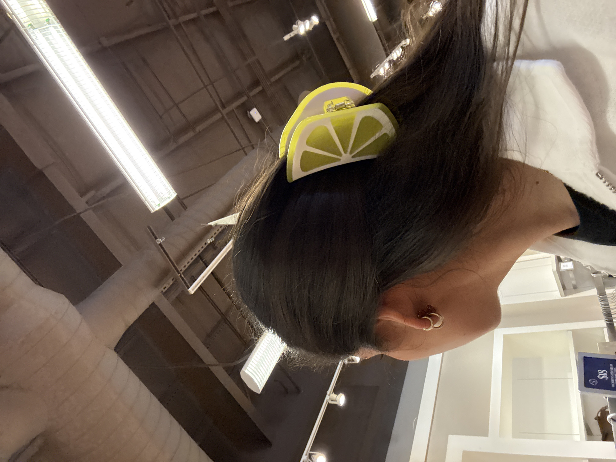 A low-angle shot depicts a person with dark hair wearing a lemon-shaped hair clip. The person also has multiple gold hoop earrings. In the background are exposed beams and florescent lights typical of a large retail establishment in the city of San Marcos, United States of America. A clothing rack and shelf unit are visible behind the person.