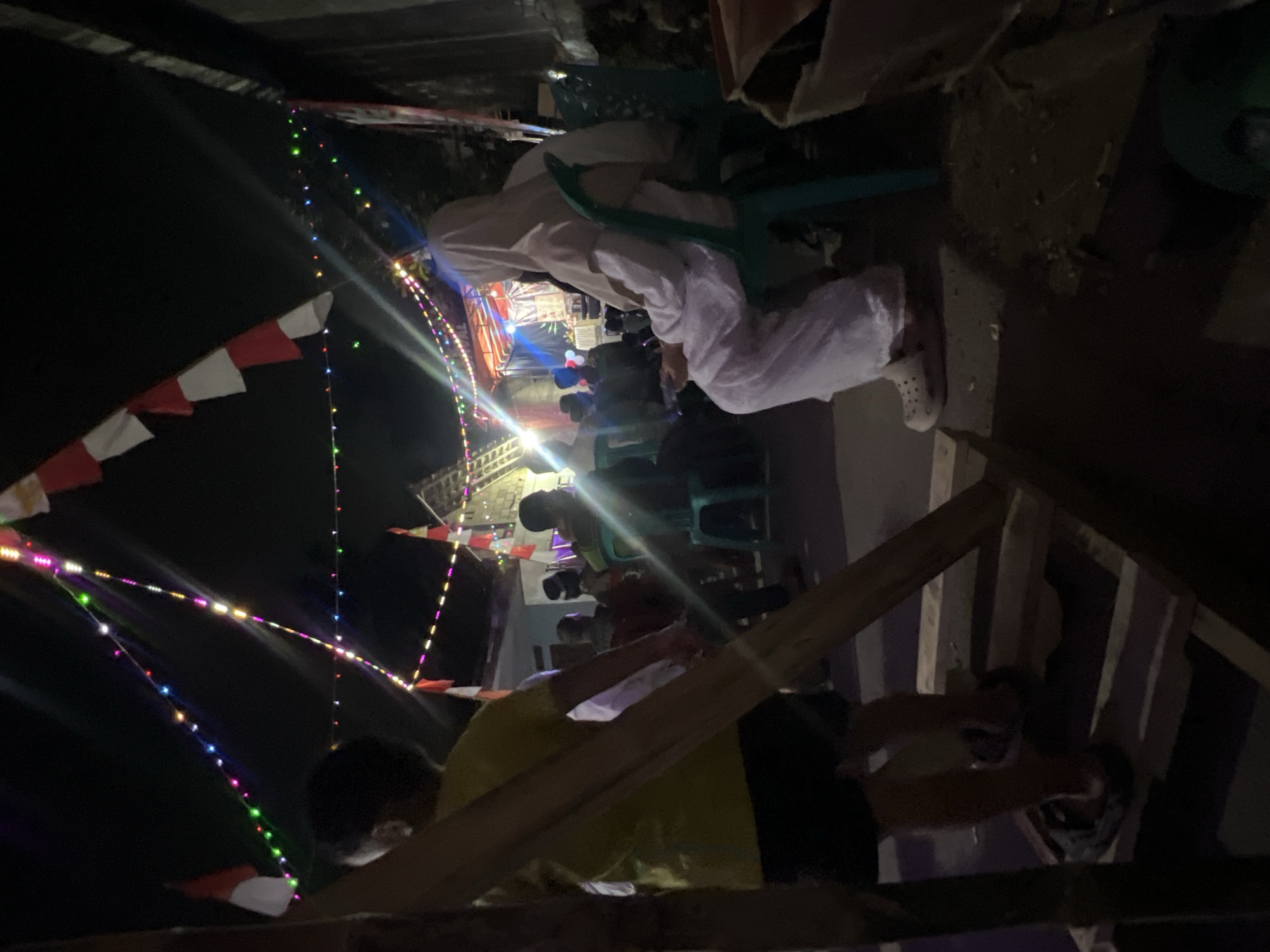 The image depicts an outdoor nighttime scene, likely in Klapanunggal, Indonesia, characterized by festive decorations. Strings of colorful LED lights are strung across the dark sky, interspersed with red and white triangular flags, suggesting a celebration or event. In the foreground, a young person in a yellow shirt and dark shorts is standing on a wooden structure, partially obscuring the view of others. To the right, a person wearing a white hijab and long white clothing is seated on a teal plastic chair, holding a mobile device. Other individuals are visible in the background, seated and standing, some under a covered stage area illuminated by stage lights. The ground appears to be a mixture of dirt and possibly pavement.