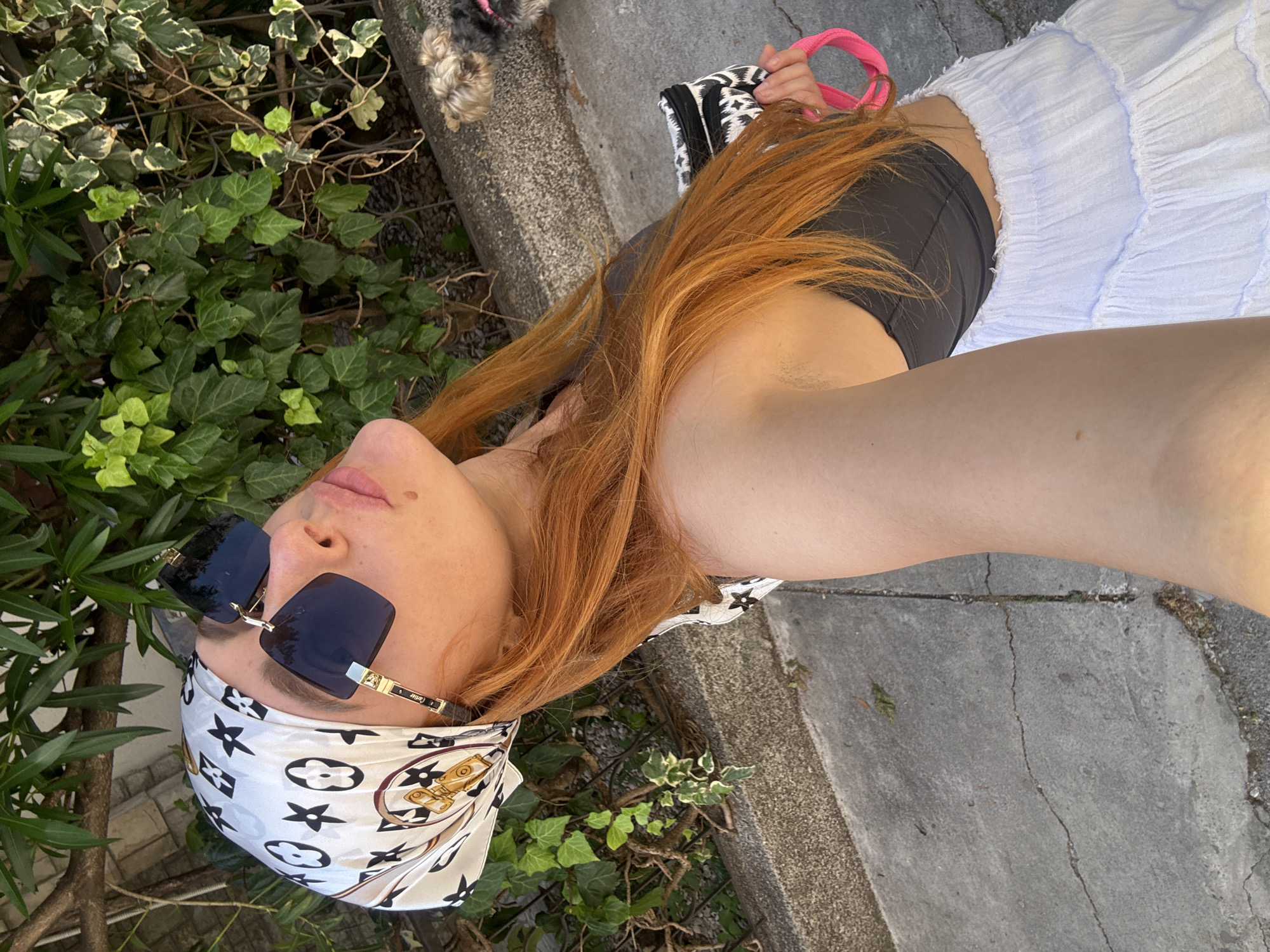 The image shows a person with long, ginger hair wearing a black top, a white skirt, and a patterned headscarf. They are also wearing large, dark sunglasses. The person is holding a small black and white patterned bag with a bright pink strap in their right hand. A small dog with light brown and grey fur is visible in the background to the right, on a leash. The scene appears to be outdoors, with green foliage and ivy growing on a fence or wall behind the person. The ground is paved with concrete. The lighting suggests it is daytime, with sunlight casting shadows. The location is identified as Budva, Montenegro.