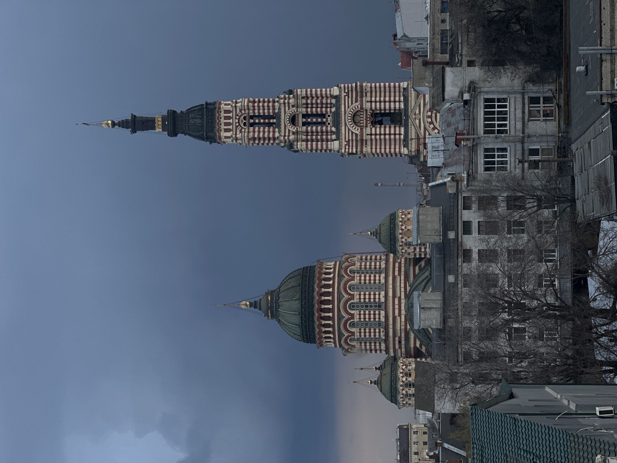 The image shows the Annunciation Cathedral and its bell tower in Kharkiv, Ukraine. The Cathedral's domes and the bell tower's striped red and white facades are prominent. The sky is overcast. Buildings surround the cathedral. Bare tree branches are visible.