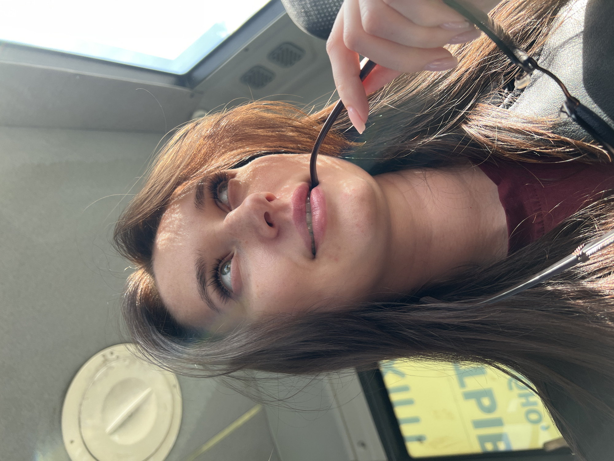 A young woman with long brown hair is inside a bus or a similar vehicle. She is holding a pair of glasses in her right hand, with one arm of the glasses in her mouth. The vehicle appears to be moving. Outside, the visible part of the window shows a glimpse of what is likely a street sign in Russian with the word "PIE." Considering the prompt's mention of Tyumen, Russia, it is inferred that the image was taken there.