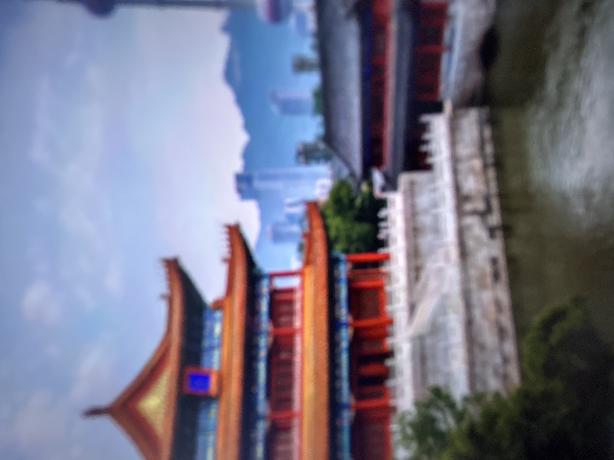 This media file displays a blurred, slightly tilted image of a traditional Chinese-style building with vibrant orange and red accents, juxtaposed against a backdrop of modern skyscrapers and a cloudy sky. The building's architectural style, characterized by its curved roofs and intricate details, suggests a cultural or historical significance. The presence of the contemporary city skyline in the background implies a fusion of old and new, possibly indicating a historical district within a modern urban environment. The water body to the right of the building and a wall structure add to the scene's composition, suggesting it might be a riverside or lakeside setting. The image is abstract due to its blurriness, making it difficult to discern specific details about the activity or time of day. No text is visible in the frame. The provided location context of Gelendzhik, Russia, is incongruent with the visual elements of the image, which strongly suggest an East Asian setting.