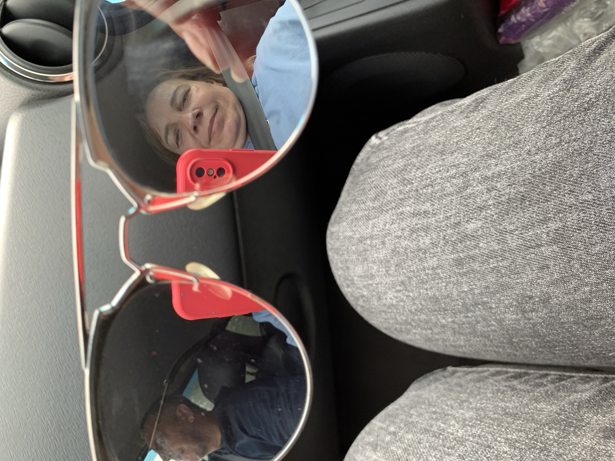 The image is a close-up, point-of-view shot taken from inside a vehicle, looking through a pair of sunglasses. The sunglasses reflect a man in the driver's seat of the car, wearing a dark blue shirt. In the reflection of the right lens, a woman is visible, wearing a light blue top and a seatbelt, holding a red smartphone. The foreground of the image shows the legs of the person taking the picture, dressed in gray jeans. The background of the image, outside the reflection, is dark and shows part of the car's interior. The location is specified as Ilyanskiy selskiy Sovet, Belarus.
