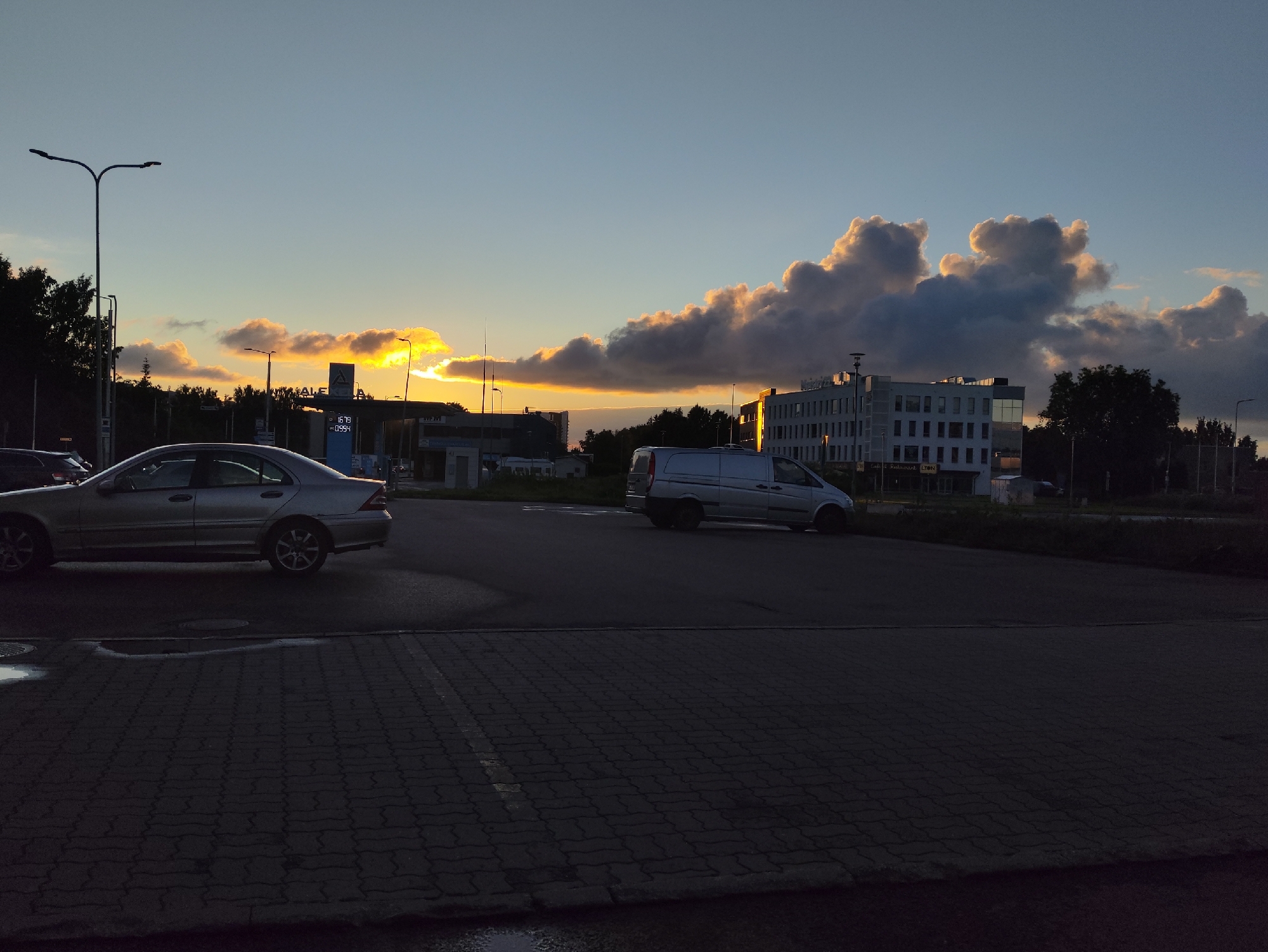 The image depicts a twilight scene in Tallinn, Estonia, likely from a parking lot. A silver sedan is parked on the left, and a silver van is visible in the middle ground. In the background, a modern, multi-story building stands, silhouetted against a sky filled with dramatic, illuminated clouds and the setting sun. A lamppost is prominent on the left side of the frame. The ground is paved with interlocking bricks. The scene suggests a commercial or office area at the end of the day.