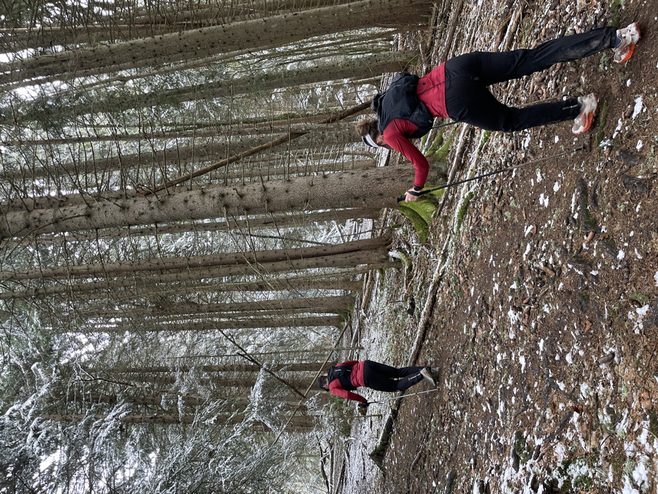 Two individuals are depicted ascending a snow-dusted, densely wooded uphill slope in a coniferous forest, identified as being in Passy, France. The ground is composed of dark soil, exposed tree roots, scattered fallen twigs, and patches of white snow. Both subjects are viewed from behind, wearing identical red long-sleeved athletic tops, black athletic pants, and black hydration vests/small backpacks. The foreground subject, positioned towards the right, leans forward, utilizing two black trekking poles planted in the ground for support. This individual wears orange and white trail running shoes and a white headband. The second subject is located further up the slope towards the left, also leaning forward and using two trekking poles. This individual wears white/light-colored trail running shoes. The forest consists of numerous tall, slender trees with dark bark, some displaying sparse snow on their branches.