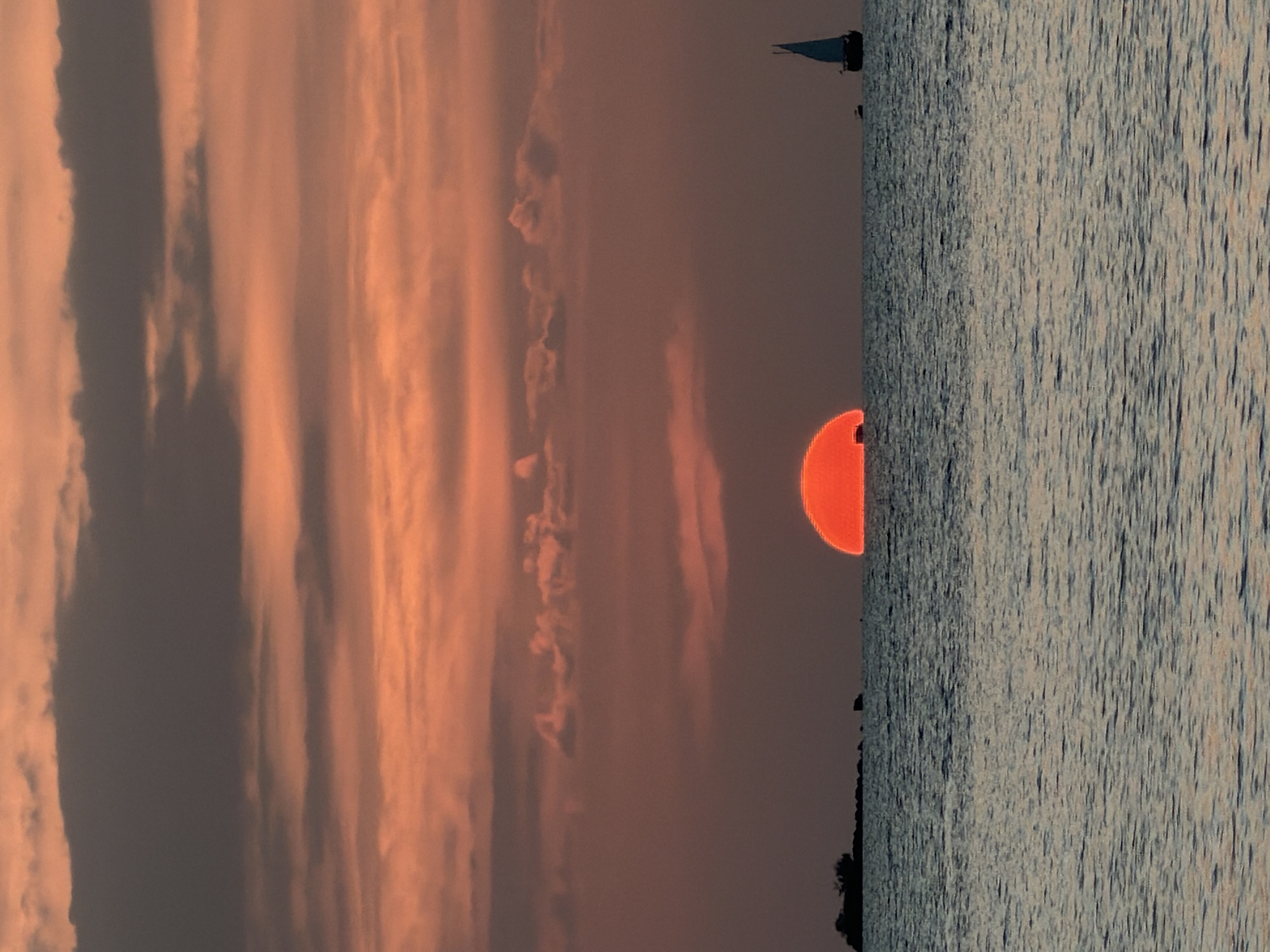 A vibrant sunset paints the sky over the ocean in Ko Samui District, Thailand. The sun, a brilliant orange orb, is partially submerged on the horizon, casting a warm glow across the clouds. The water below reflects the sky's colors, with gentle waves creating a textured surface.
In the distance, a small island with sparse vegetation stands silhouetted against the horizon. A sailboat with its mast and sail visible navigates the sea to the right of the frame. Two smaller boats are also present in the distance, one near the island and another closer to the sailboat. The scene is tranquil and serene, capturing the natural beauty of a coastal sunset.