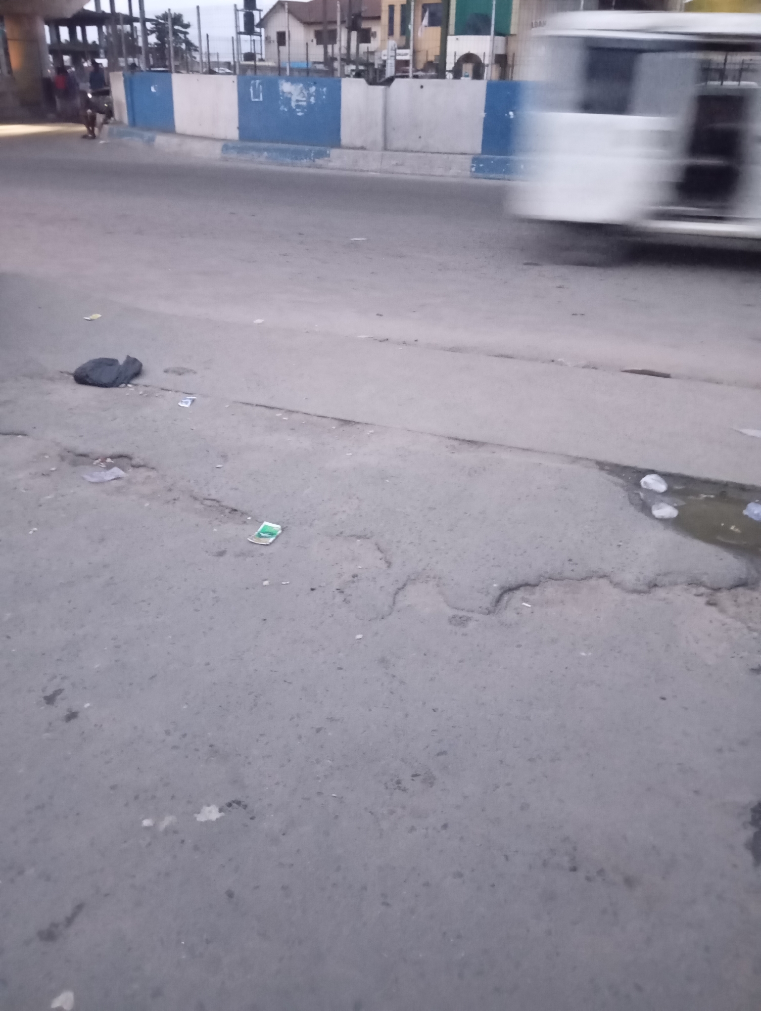 The image shows a section of an urban road in Port Harcourt, Nigeria, likely near an overpass or elevated roadway, given the structure visible in the background. The road surface is made of asphalt, showing signs of wear and tear with several small potholes and cracks. To the left, a black trash bag lies on the road. In the middle ground, there are scattered pieces of litter, including what appears to be a crumpled green and white wrapper and some small paper scraps. A pool of murky water with floating debris is visible on the right side of the road. In the background, a blue and white striped barrier runs along the side of the elevated roadway. A white bus is captured in motion, blurred due to movement, indicating traffic. A few indistinct figures and what seems to be a motorcycle are also present near the barrier, suggesting general street activity. The overall scene depicts a typical urban roadside environment with elements of urban decay and ongoing traffic.