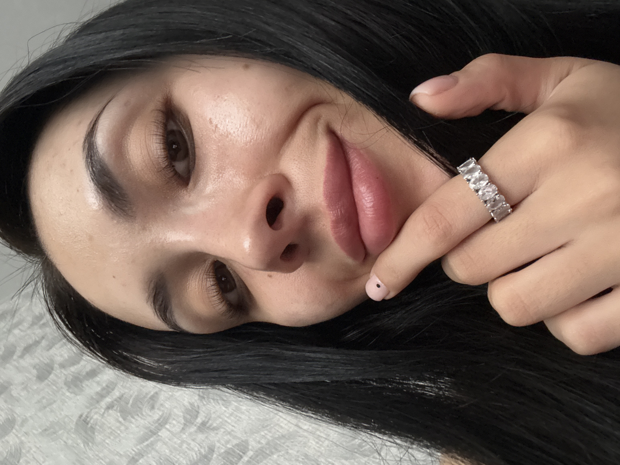 A close-up, slightly angled view of a woman's face with her dark hair framing her face. She is wearing a silver ring with baguette-cut stones on her finger which is near her lips. Her skin appears clear with a slight sheen. She has full lips. One fingernail is painted pink with a black dot. The background is a textured white surface. The image shows no indication of the location being Votkinsk, Russia.