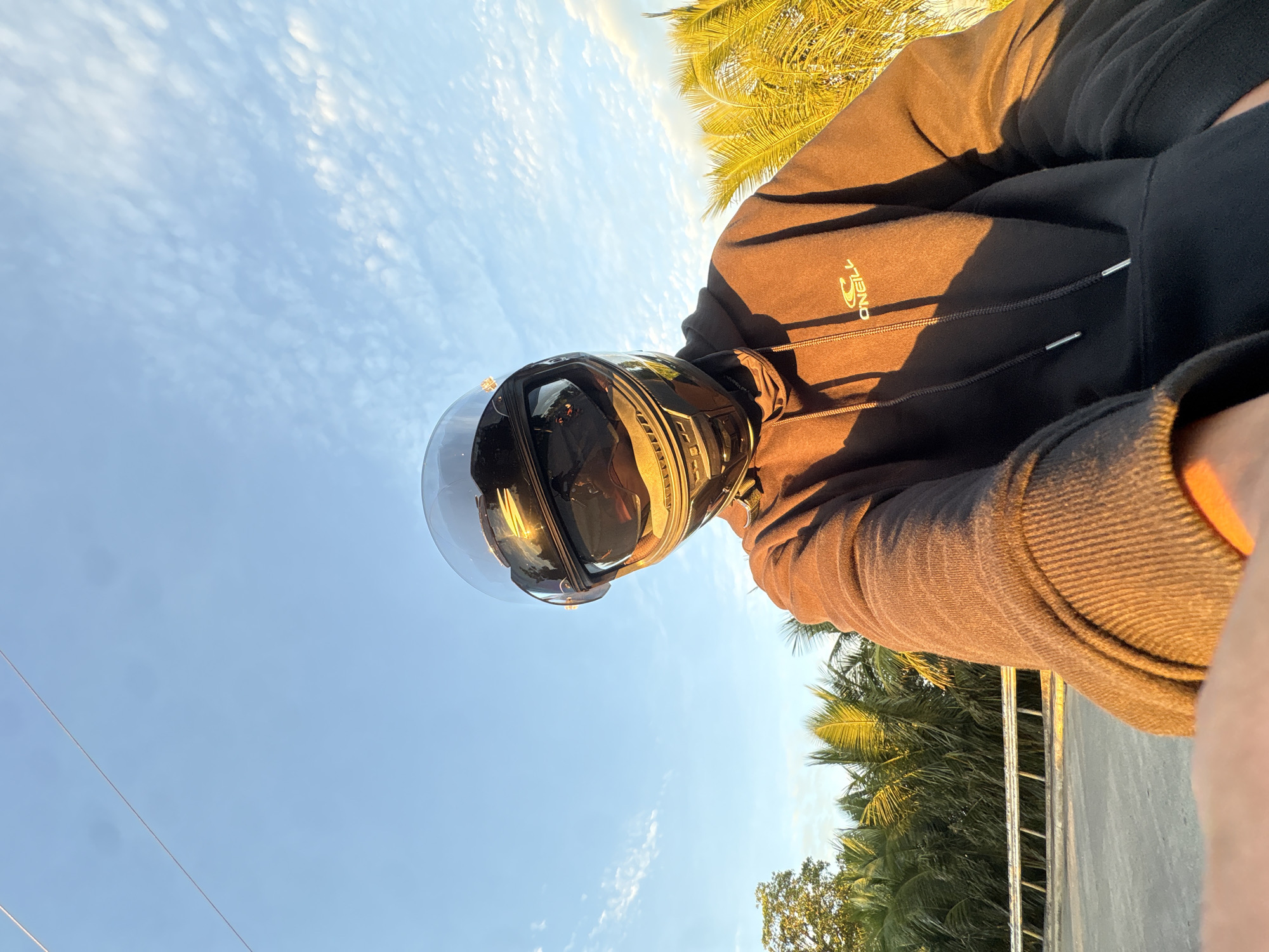 A person wearing a black full-face motorcycle helmet with a reflective visor and a brown hooded sweatshirt is taking a selfie outdoors during golden hour. The person is partially visible, with the helmet and upper body in focus. The background features a clear blue sky with scattered white clouds, a line of palm trees, and a road with a guardrail. The lighting suggests late afternoon or early morning, casting a warm glow on the scene. The location is identified as Davao City, Philippines.