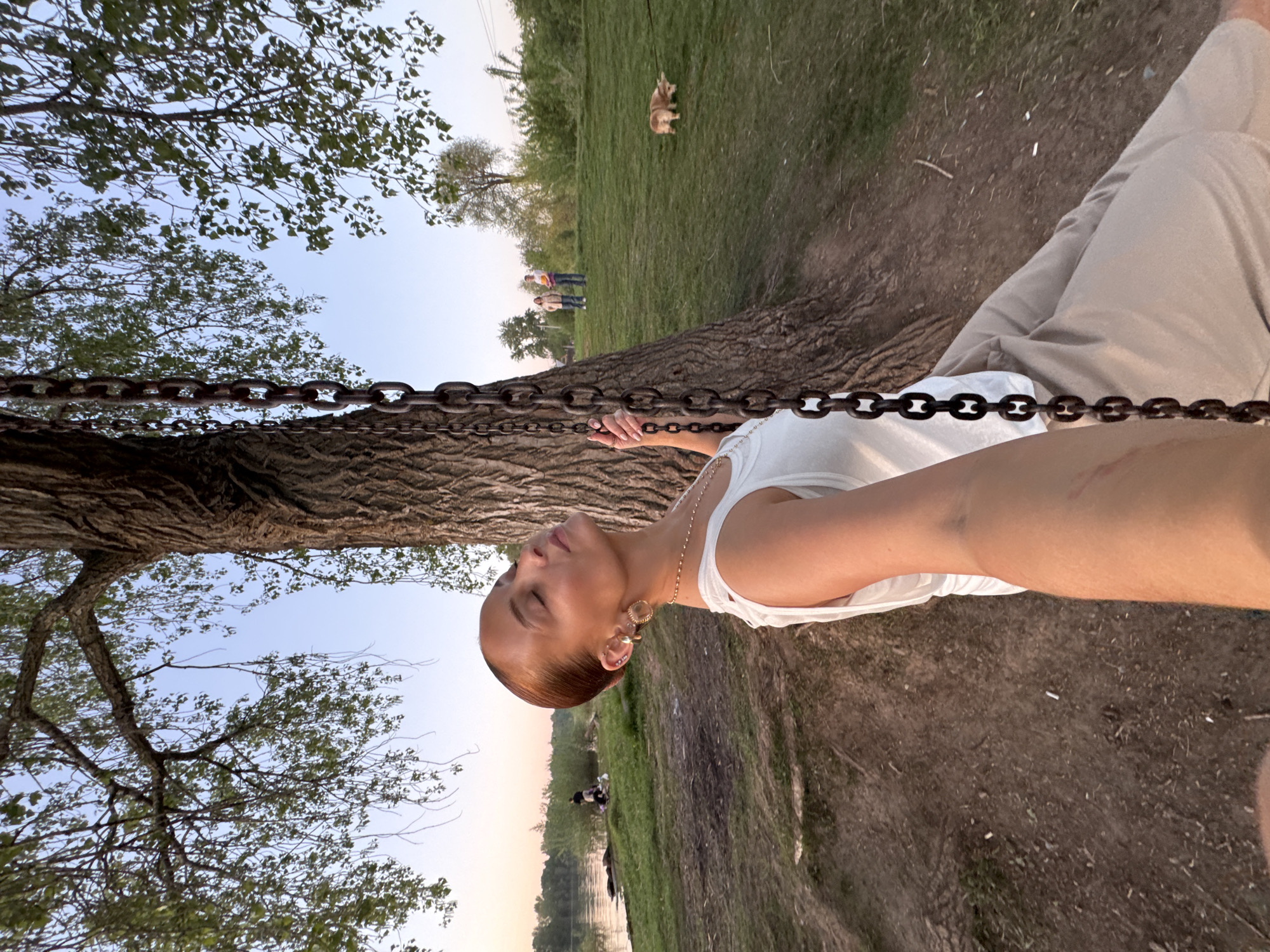 A person, likely female, is holding a chain attached to a tree. The location appears to be a park or green space near a body of water, potentially in or near Chaikovsky, Russia, based on the request. In the background, there are figures of other people and a dog visible in the distance. The environment includes grass, trees, and a water body. The sky is partially visible through the tree branches.