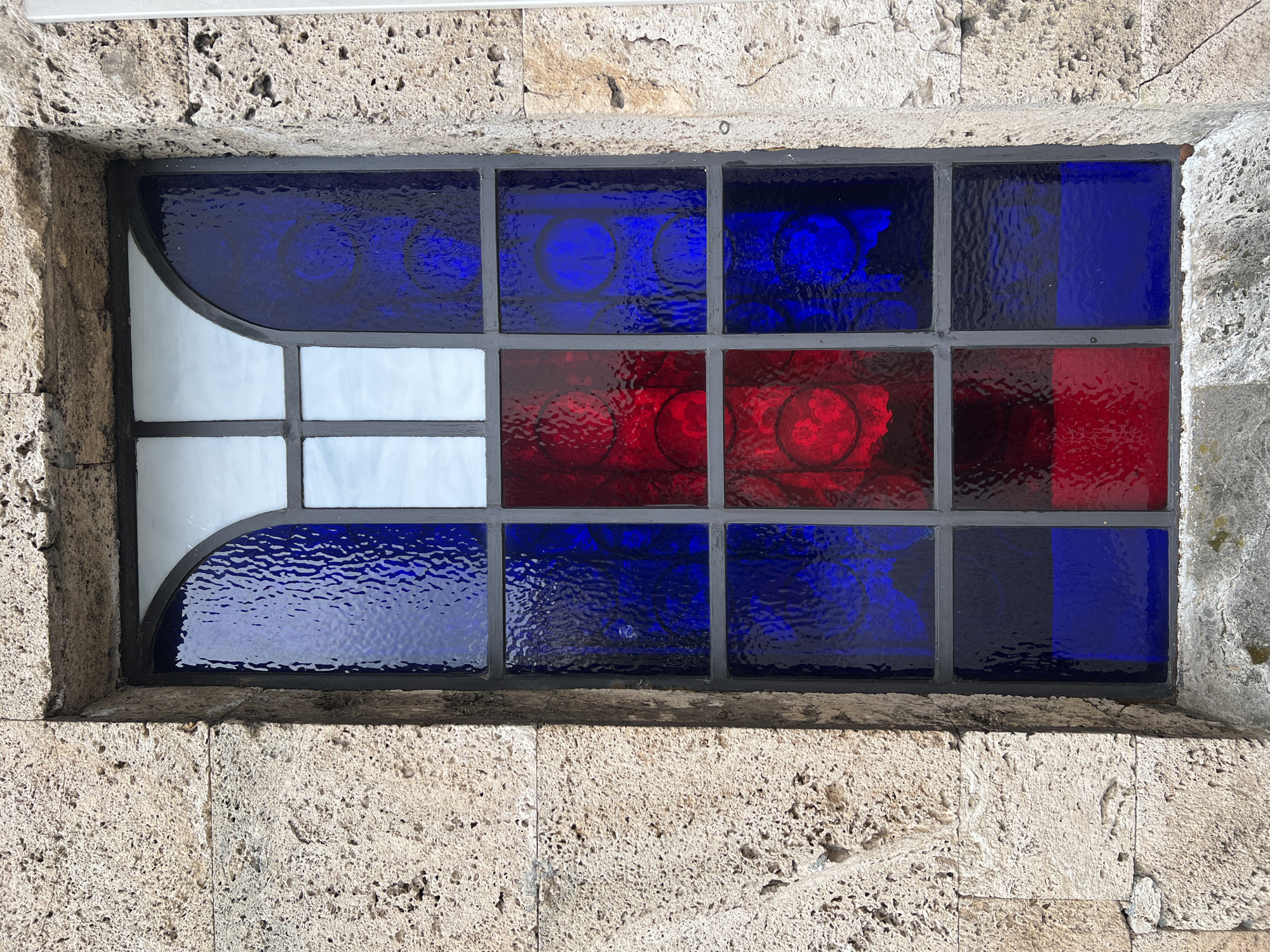 A rectangular stained glass window is set within a stone wall. The window consists of 12 panes separated by dark grey mullions and frames. The panes are arranged in a 3x4 grid. The upper row is blue, the middle row is red, and the bottom row is blue. The pane on the left-most side consists of white panes and a curved blue pane. The red panes feature circular patterns. The stone wall is textured and made of light beige-colored blocks. The photo was likely taken in Burgas, Bulgaria.