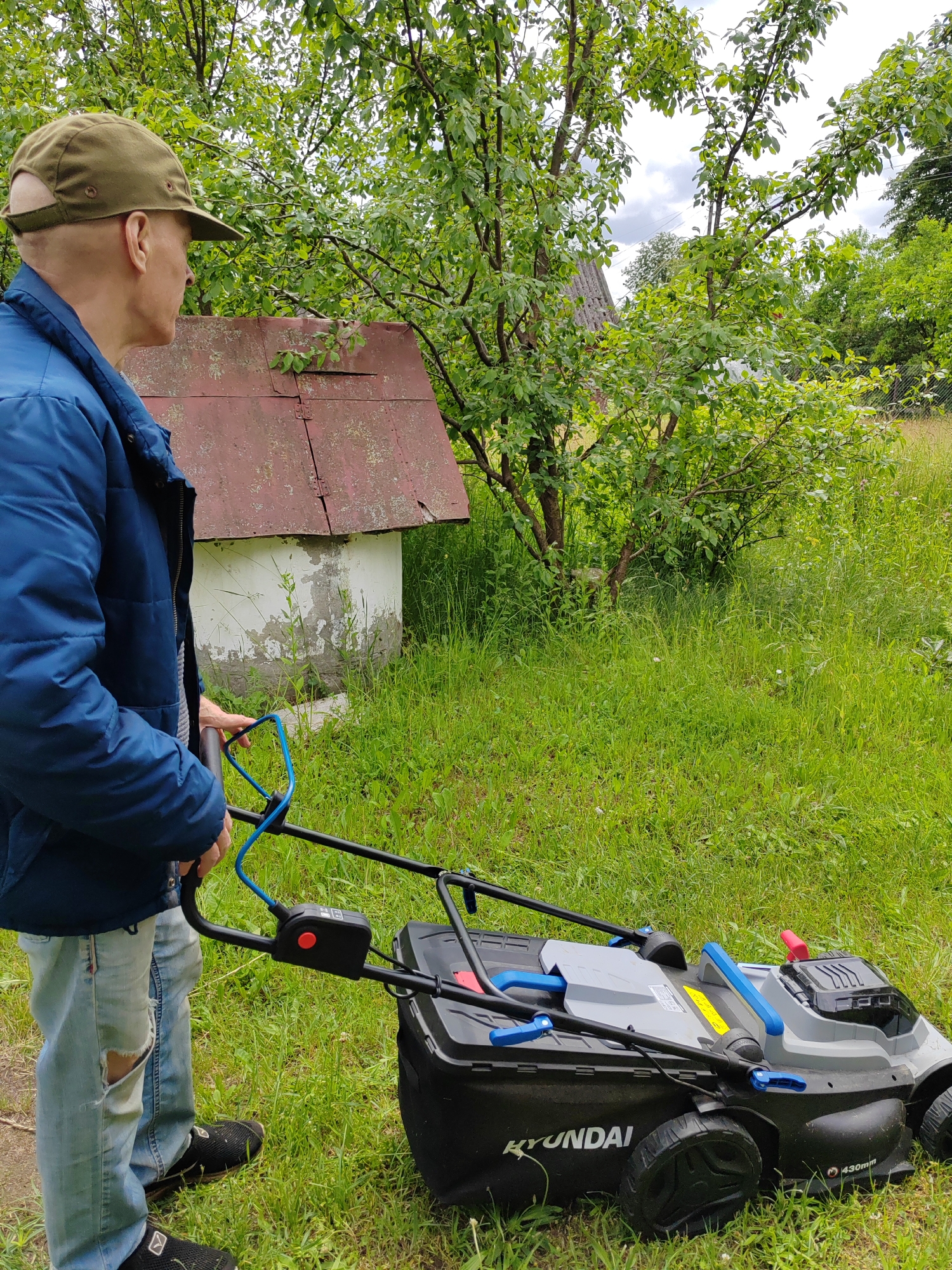 A man wearing a blue jacket, a green cap, and ripped jeans stands in a grassy area, holding the handle of a grey and black Hyundai lawnmower. The lawnmower has a black grass bag attached. Behind the man, a circular well with a weathered red roof is visible. Trees with green leaves surround the scene, and the ground is covered in tall green grass. The sky is overcast with clouds. The location appears to be a rural area.