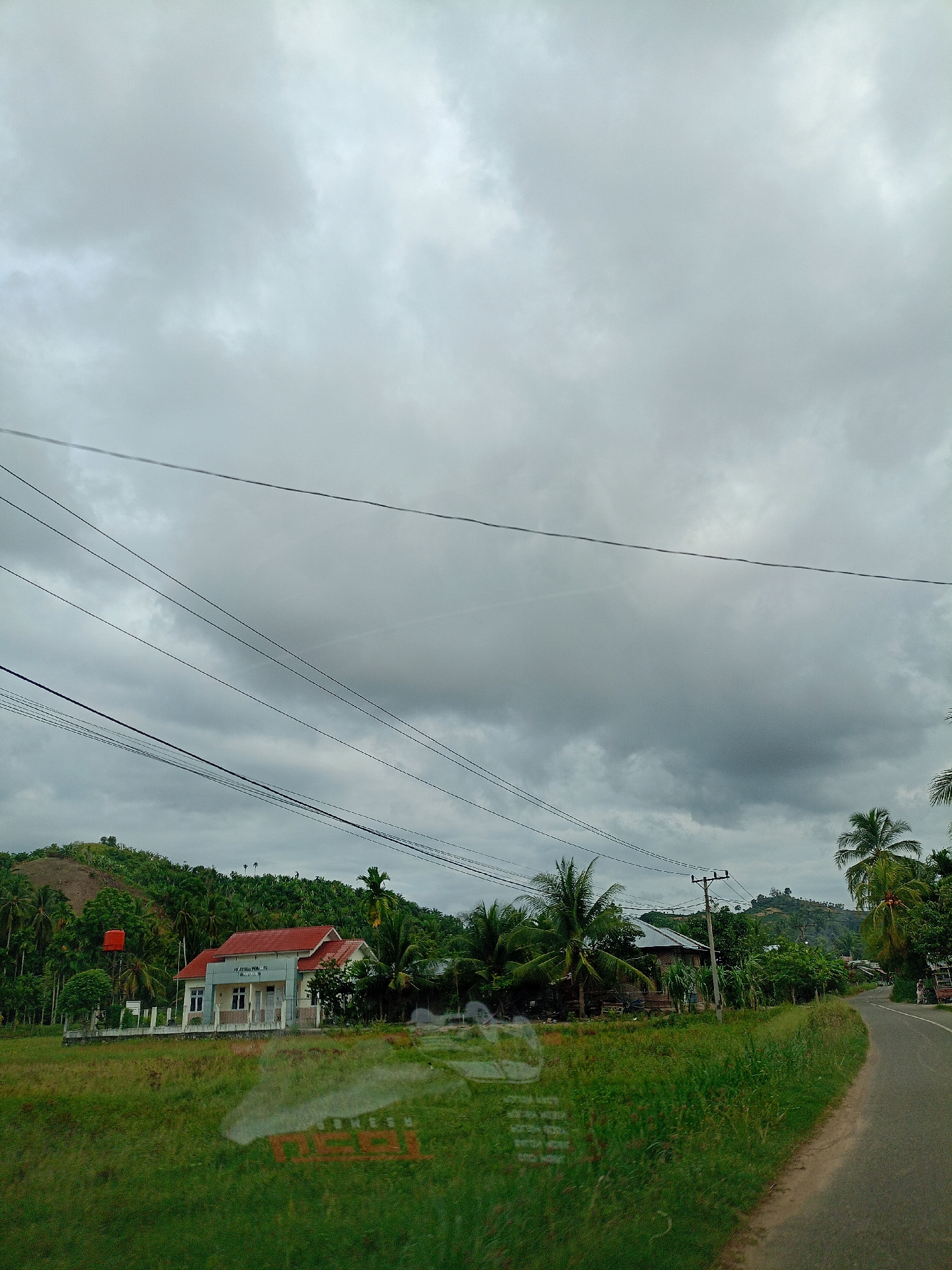 The image depicts a rural area near Palohpanyang, Indonesia. The sky is overcast with gray clouds, casting a diffuse light across the scene. A paved road curves into the distance on the right. Lush green grass and vegetation fill the foreground and separate the road from the built environment. On the left side of the image, there is a small building with a red roof, identified as a government building (Kantor Desa). Behind this building is a low hill covered with trees and greenery. Power lines are visible stretching across the sky. Palm trees are scattered throughout the landscape. In the middle distance, another house is partially visible amongst the vegetation. The camera appears to be positioned inside a vehicle. The scene is static and rural.