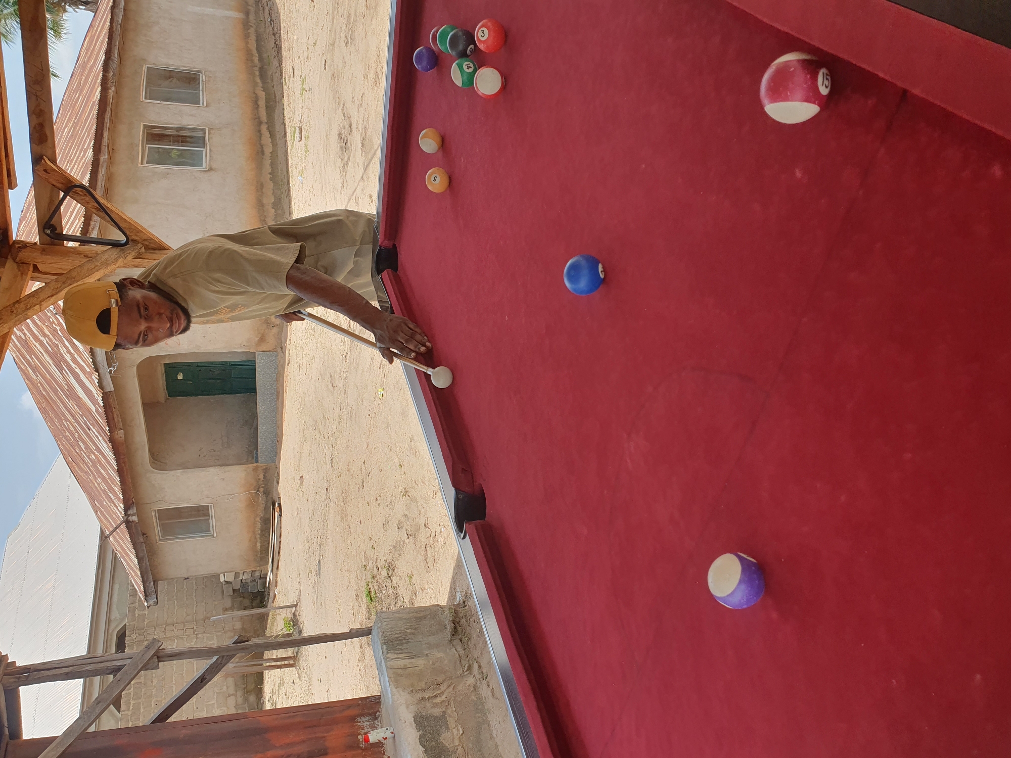 An outdoor scene in Lokoja, Nigeria, shows a man playing billiards on a red felt table. He is wearing a tan t-shirt and a yellow baseball cap. He is holding a pool cue, aiming at a white cue ball. Several numbered pool balls are scattered on the table. In the background, there are simple buildings with corrugated metal roofs. The ground is sandy.
