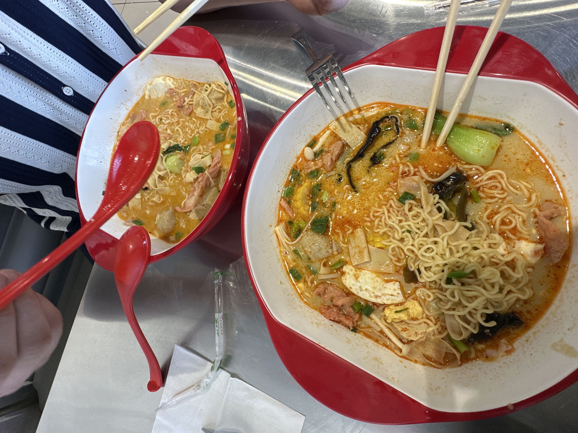 Two bowls of noodles are on a metallic table. The bowls are white with red rims. One bowl contains noodles, broth, vegetables, and meat. A pair of wooden chopsticks are sticking out of it, along with a fork. The other bowl contains a similar mixture, and a person wearing a striped shirt is holding a red spoon in it. There is another red spoon next to the bowl, along with some napkins. The scene appears to be inside a restaurant. Blagoveshchensk, Russia is not identifiable from this image.