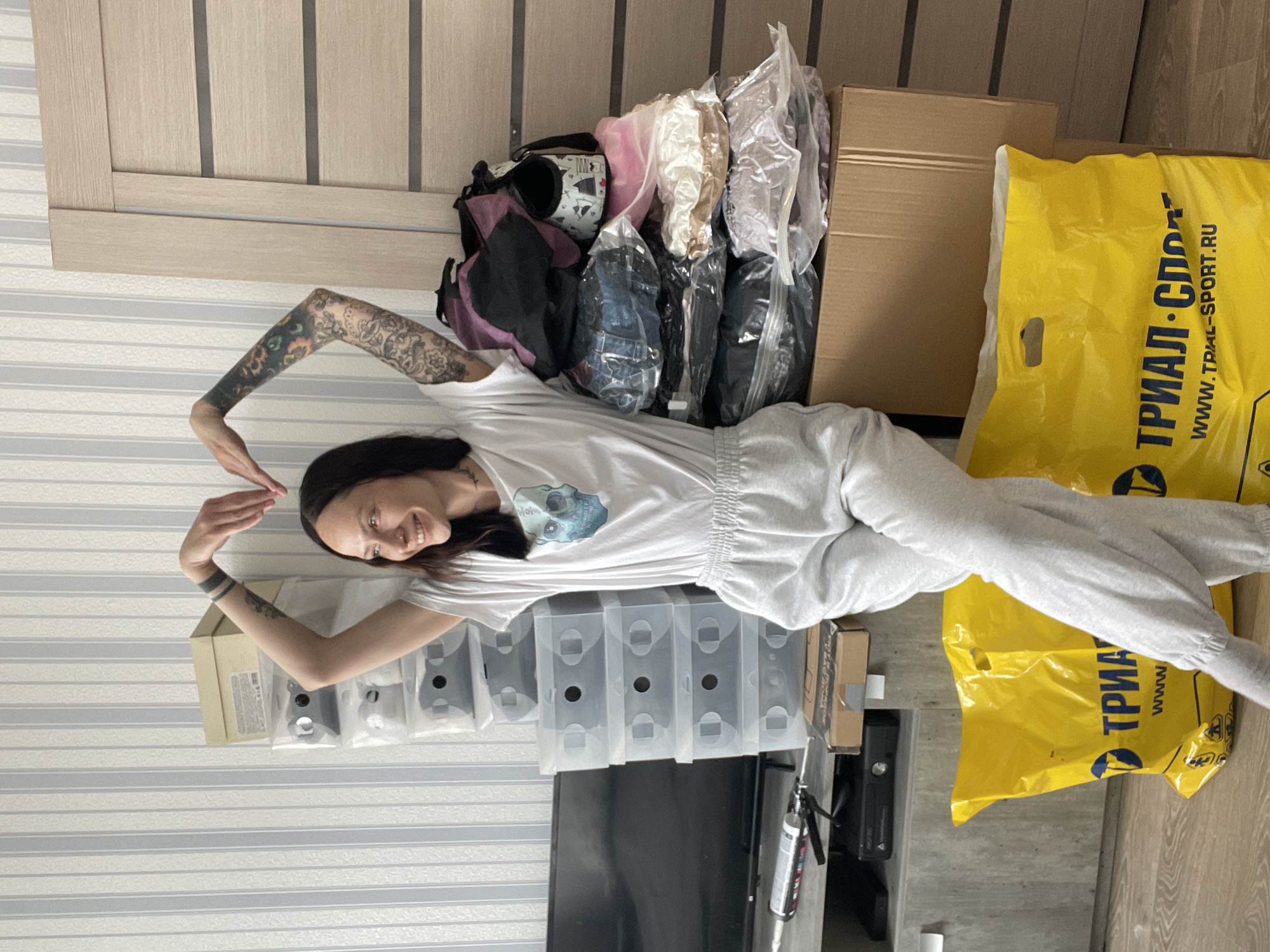 A person lies on the floor in a room. The person is smiling and making a heart shape with their hands. They are wearing a white t-shirt and gray sweatpants. The person has tattoos on their arms. Behind the person is a stack of objects, including a backpack, clear bags, a cardboard box, and a yellow plastic bag with the text "ТРИАЛ-СПОРТ" and "WWW.TRIAL-SPORT.RU". Behind the objects is a door with vertical panels. To the left of the person is a stack of clear plastic boxes and a black television. The wall is striped. The location is potentially in Izhevsk, Russia, based on the text on the bag.