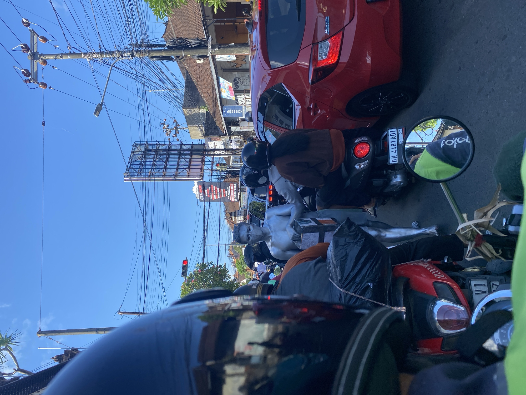 The image, taken from the perspective of a motorcycle rider in North Kuta, Indonesia, shows a congested street scene under a bright, clear sky. Several motorcycles are lined up in traffic, with riders wearing helmets. A red Honda car is positioned to the right. Overhead, a dense network of power lines crisscrosses above buildings and billboards. A silver-painted person sits as a statue on a motorcycle.