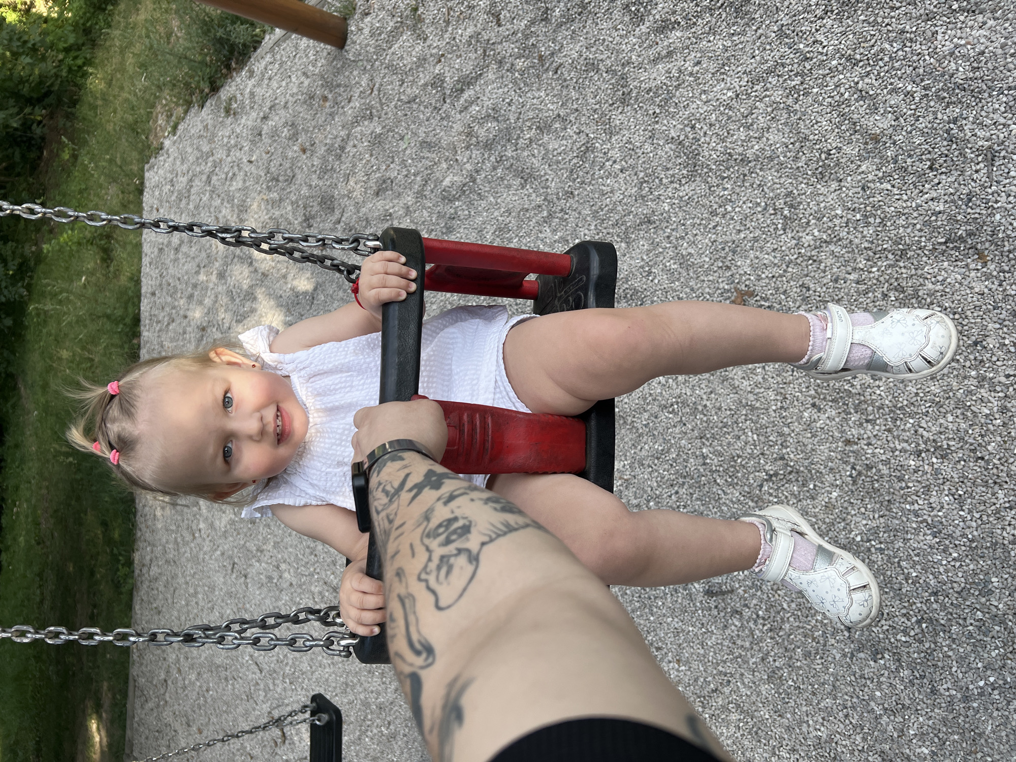 The image shows a toddler sitting on a swing at a playground. The toddler, a female, is wearing a white dress and white sandals with pink socks. She has blonde hair in pigtails. A tattooed arm is visible holding onto the swing's safety bar, suggesting an adult is present and interacting with the child. The playground surface is covered in gravel, and trees and foliage are visible in the background. The scene appears to be outdoors during daylight hours.