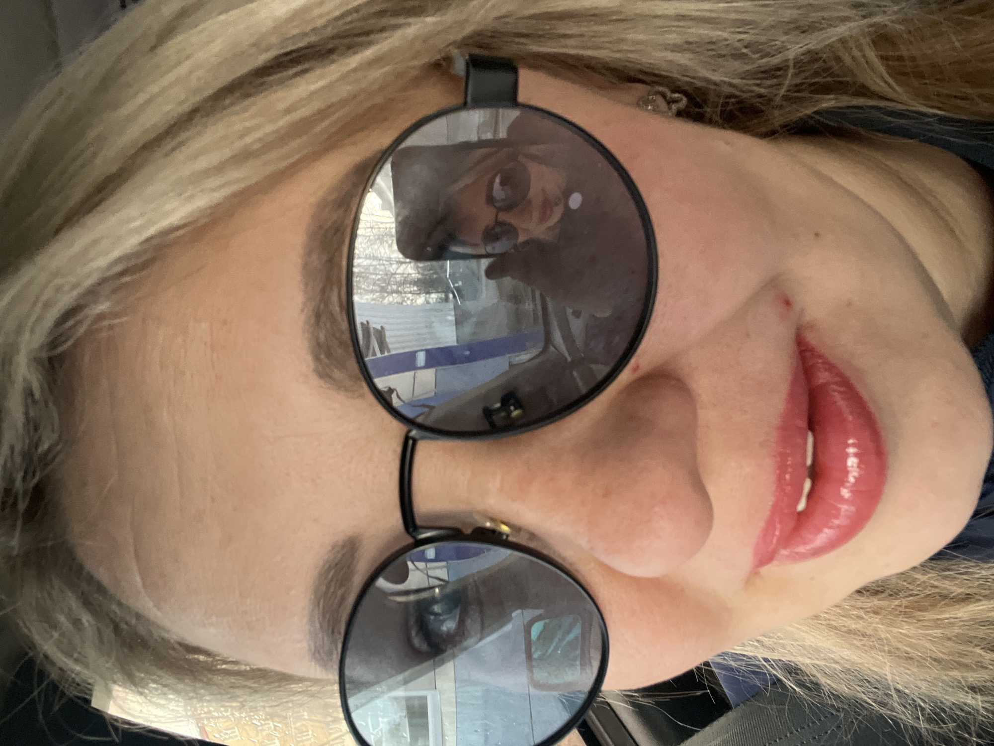 A close-up selfie captures a person, likely female, with blonde hair and fair skin. The subject is wearing large, circular sunglasses with dark lenses and black frames, which dominate the upper part of the face.
A clear reflection in the upper lens of the sunglasses reveals the person holding a smartphone, confirming the image is a selfie. The reflection also provides a glimpse of the surrounding environment, showing what appears to be an indoor space with windows, suggesting the subject might be inside a vehicle or building. Elements such as fence-like structures and distant buildings are faintly visible through the reflected windows.
The person has well-defined eyebrows and is wearing glossy pink lipstick. A small reddish mark is present near the right corner of the mouth. A small, metallic earring is visible on the subject's right earlobe. The image is tilted, presenting the face at an angle. No specific activity beyond taking the photo is depicted, and no text is visible in the frame. The location context is Chaikovsky, Russia.