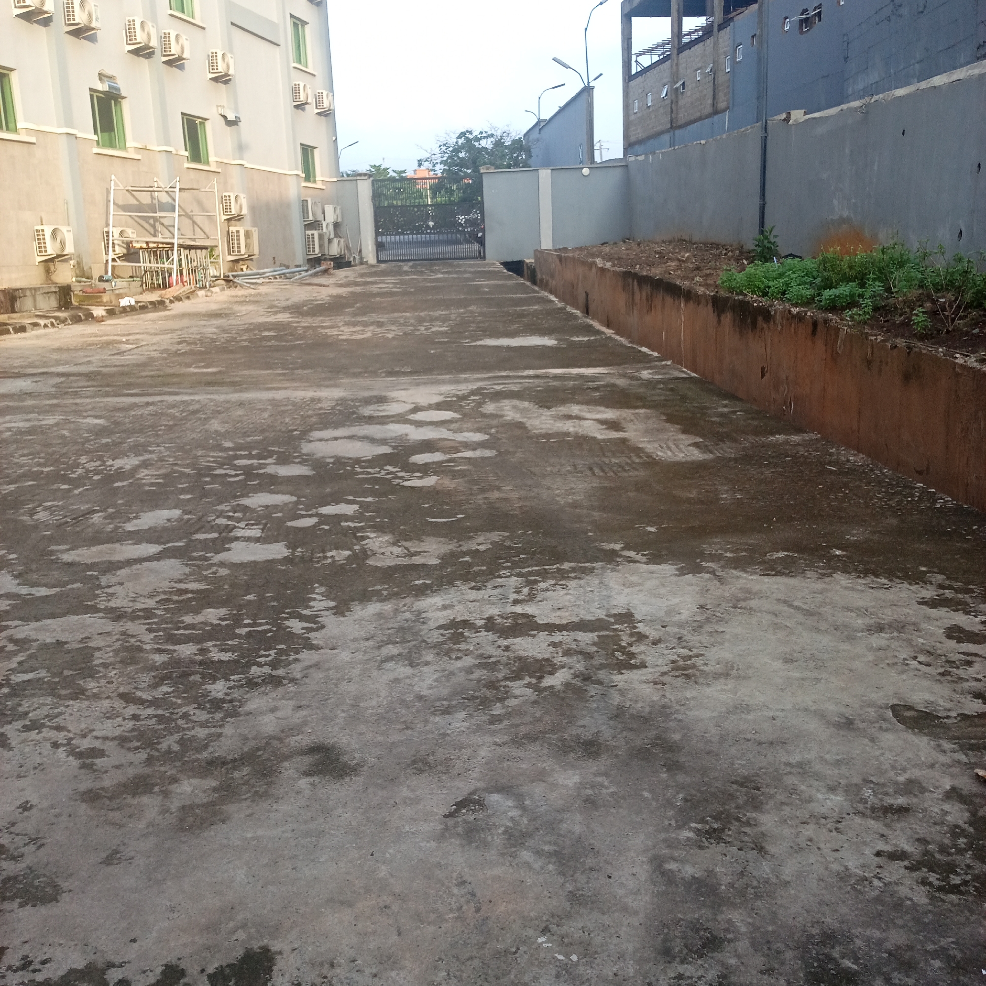 The image captures an outdoor scene in Ibadan, Nigeria, featuring a paved area leading towards a gated entrance. To the left, a light-colored, multi-story building is visible, adorned with numerous air conditioning units mounted on its exterior. Some of these units are clustered together, while others are individually placed. A metal scaffolding structure is also present near the building's base. The paved ground in the foreground exhibits a weathered and mottled appearance, with patches of lighter and darker concrete, suggesting wear and possibly moisture.
Beyond the paved area, a metal gate stands at the entrance to what appears to be a compound or driveway. Streetlights are positioned on poles, with one extending over the gate area. A grey wall runs along the right side of the image, delineating the boundary of the property. Beyond this wall, a strip of brown earth is visible, with some sparse green vegetation growing along its edge. In the background, a large, dark grey building is partially visible, with a concrete structure rising above it. The sky above is overcast, indicating a cloudy day. The overall impression is one of an urban setting in a state of development or possibly maintenance. There are no people or active interactions visible in the image.