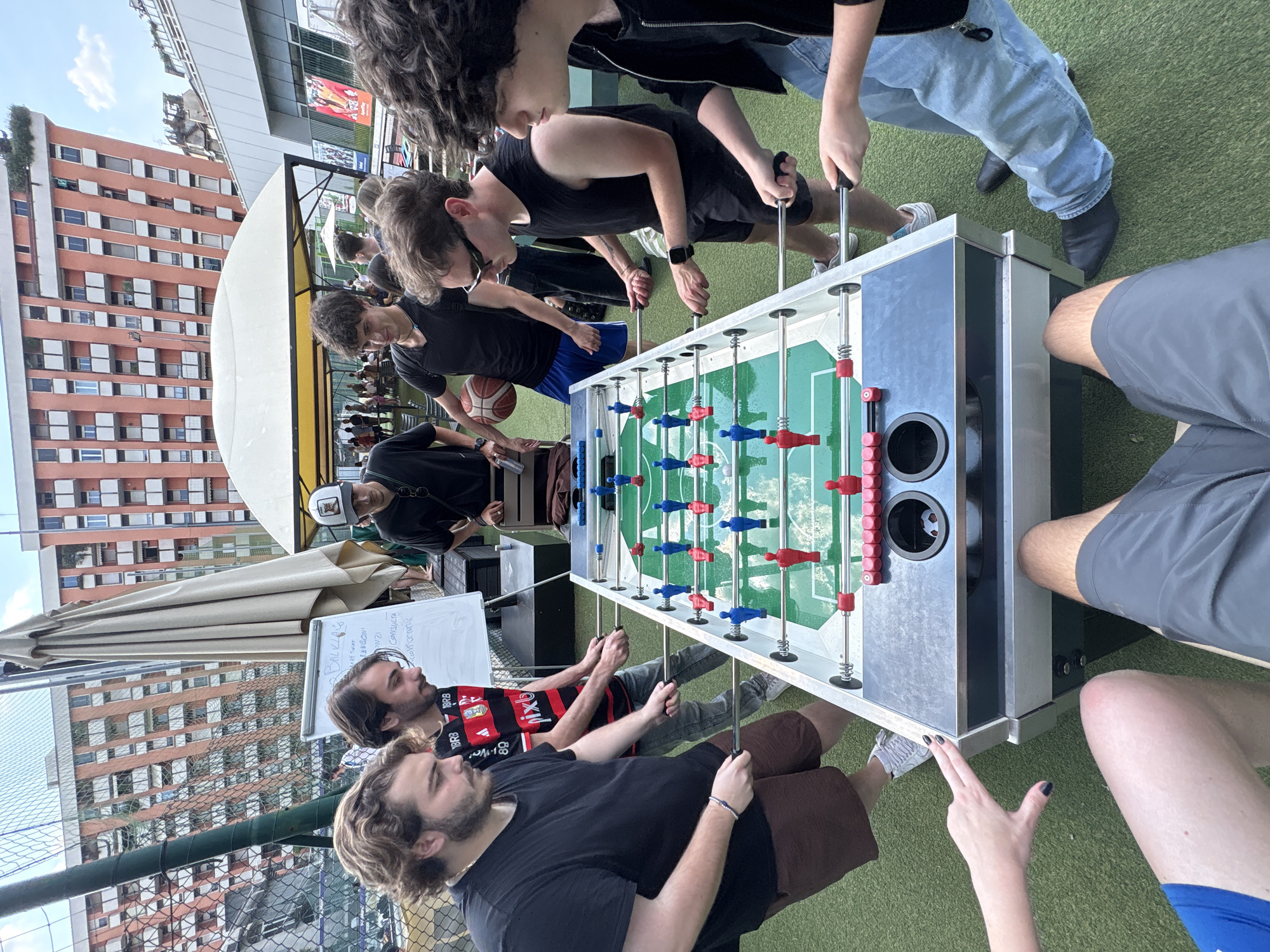 An elevated outdoor setting in Milan, Italy, features a group of individuals engaged in or observing a foosball game on a green artificial turf surface. Six individuals are actively positioned around a multi-colored foosball table with red and blue players, gripping the metal rods.
On the left side of the table, one man with curly brown hair and a beard, wearing a black t-shirt and brown shorts, holds two sets of rods. Adjacent to him, another man with a beard and brown hair, wearing a red and black striped t-shirt with "BRB" and "pixl" logos, also operates the rods. To their left, a chain-link fence is visible.
On the right side of the table, a man in a black sleeveless top, black shorts, and sunglasses manipulates the rods. Next to him, a man with dark curly hair, wearing a black long-sleeved shirt and light blue jeans, is also holding rods. Below the table, two pairs of legs belonging to players are visible, one in blue shorts and another in grey shorts. A female hand with dark-painted fingernails extends from the left foreground towards the table's edge.
Behind the players, several individuals are present. One man wearing a black t-shirt and blue shorts holds a basketball under his arm. Further back, under a white awning or tent, a gathering of people is visible. A person wearing a white baseball cap with an animal logo stands near a white easel-mounted whiteboard displaying handwritten text, including "BALKLAS," "ALENCONI," "21," "CONSULTA," and "OCTOPUS."
The background is dominated by multi-story residential buildings with orange-toned facades, numerous windows, and balconies, typical of an urban environment. Some rooftops show vegetation. A large, folded beige umbrella is positioned near the white tent. The scene is illuminated by daylight under a clear sky.