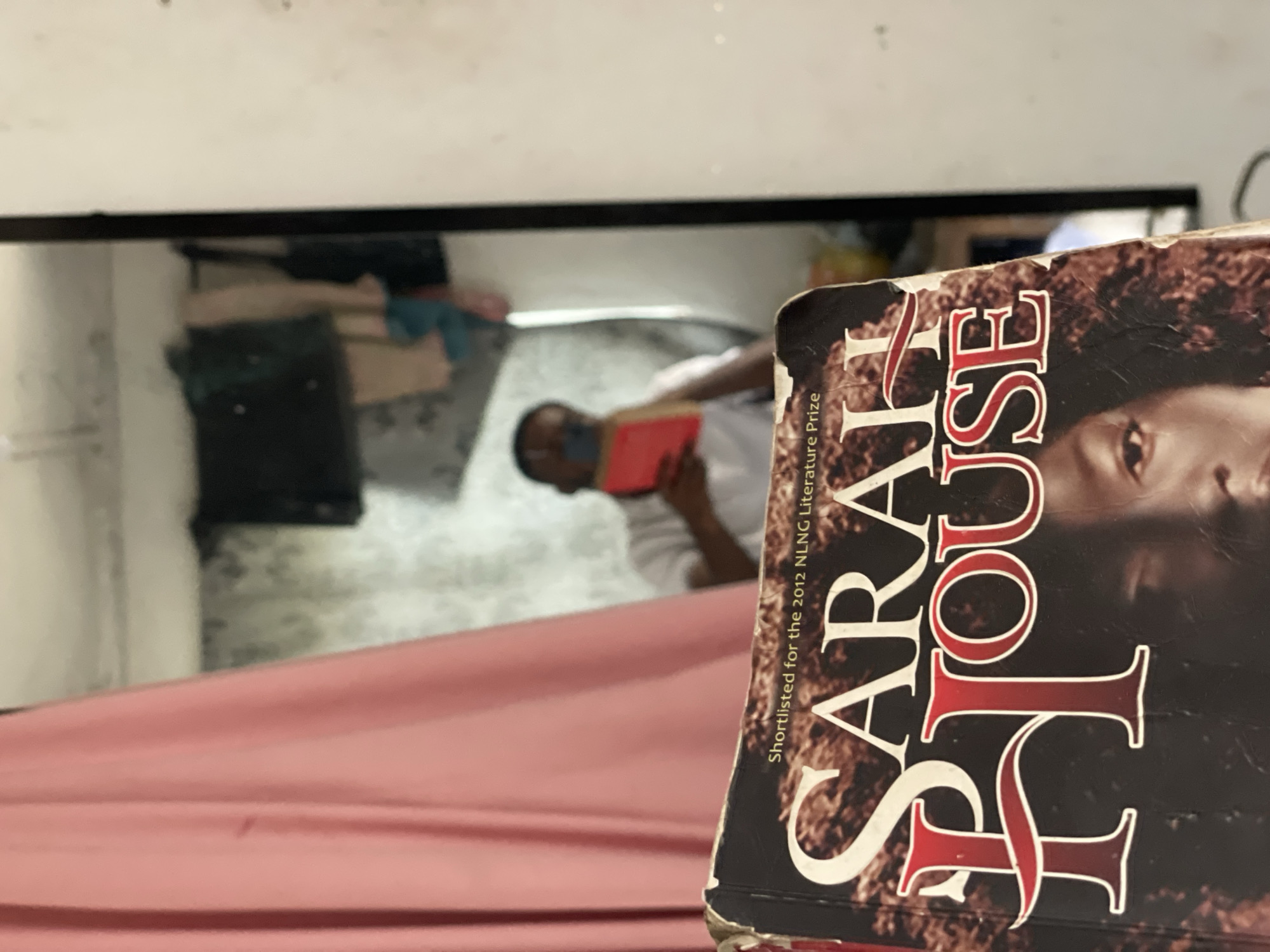 A person lies on their back in a bed, face obscured by a book titled "Sarah House," the book's cover facing the viewer. The person is holding a red book or object in their hands. The scene is reflected in a mirror above the bed. The background suggests an interior space, possibly a room. The location is not discernible as Lokoja, Nigeria, from the given image.