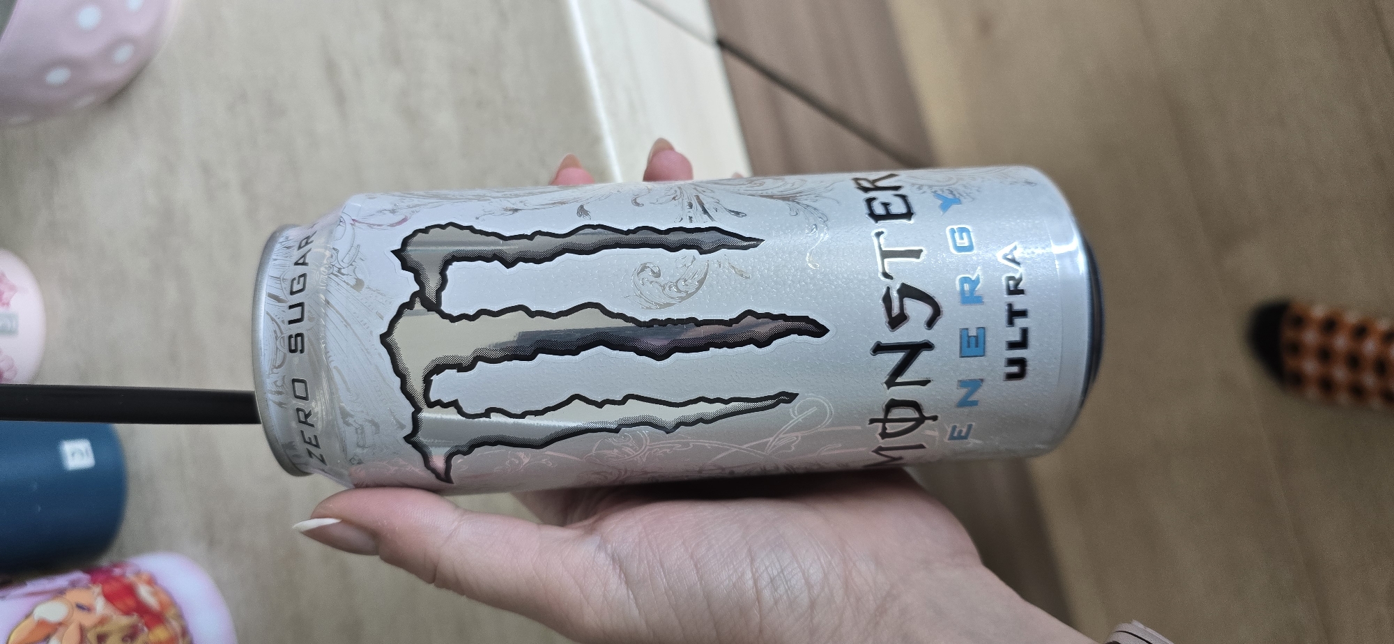 A close-up, slightly rotated image captures a person's hand holding a Monster Energy Ultra Zero Sugar drink can with a black straw inserted. The hand, likely female, has well-kept nails with a light polish.
The energy drink can is predominantly silver-white with a textured appearance and features intricate, subtle silver or light grey decorative patterns. The iconic Monster logo, a jagged "M," is prominent in silver with a dark outline. Visible text on the can includes "MONSTER ENERGY" in bold, with "ENERGY" being smaller and colored blue. Below this, "ULTRA" is also displayed in blue text. Along the side of the can, the words "ZERO SUGAR" are printed vertically.
The background suggests an indoor setting, with a light-colored surface resembling a countertop or table visible above the can. Below the hand, a wooden floor is discernible. Various other objects are partially visible in the background: a pink bowl with white polka dots at the top left, a small blue item (possibly another cup or container) at the bottom left, and a pink object featuring a cartoon character further to the bottom left. In the upper right background, part of a foot wearing an orange and black patterned sock is visible. The lighting is ambient, indicating an indoor environment, but the specific time of day is not evident. No particular emotions are discernible from the visible hand. The overall scene suggests a casual moment, perhaps taking a break, in a home or similar indoor space.