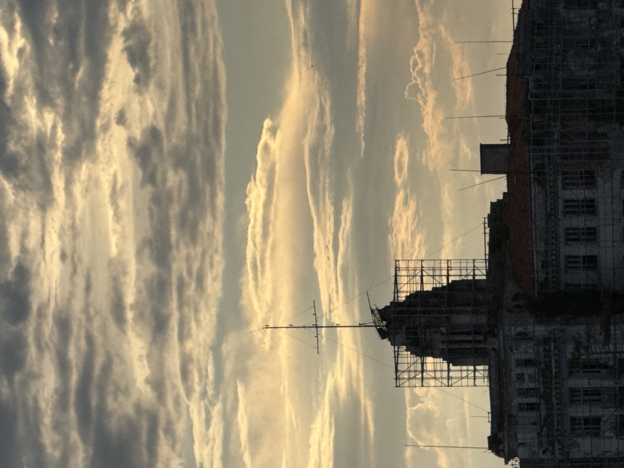 The image depicts a building under construction or renovation in Sozopol, Bulgaria, viewed against a backdrop of a cloudy sky at sunset or sunrise. The building's facade is obscured by scaffolding, suggesting ongoing work. An antenna or similar structure is mounted on top of the building. The sky is filled with elongated clouds illuminated by the sun's golden light. The perspective is from a low angle, tilting upwards towards the sky. The scene conveys a sense of transition, with the building in progress and the sky displaying vibrant colors.