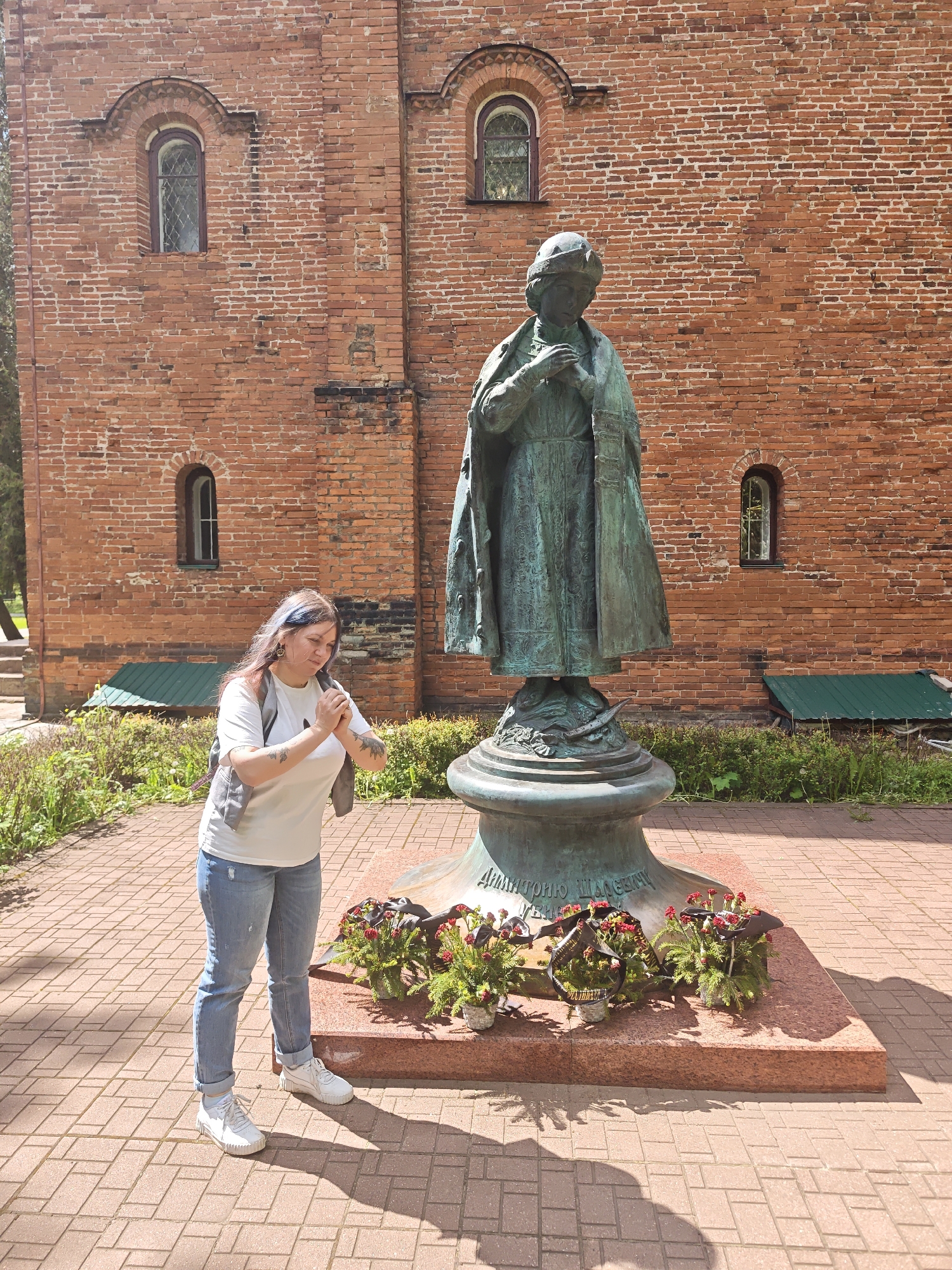 A full outdoor shot captures a woman and a statue set against a red brick building. The woman, positioned on the left, has fair skin, light blue hair streaks, and is wearing a white t-shirt, light blue jeans, a gray vest, and white sneakers. Her hands are clasped together, held up near her chest as she stands close to the statue. The bronze statue depicts a figure in a long robe, standing on a pedestal. The base of the pedestal is surrounded by small floral wreaths. The statue and woman are positioned on a brick-paved area. Behind the statue, the red brick building has several small, arched windows. Green plants and a hint of trees are visible on the far left of the image. The scene is well-lit with sunlight, casting shadows on the brick pavement. The words "Дмигторио Шассеми" are visible on the statue base. The location is in the city of Unknown, Unknown.