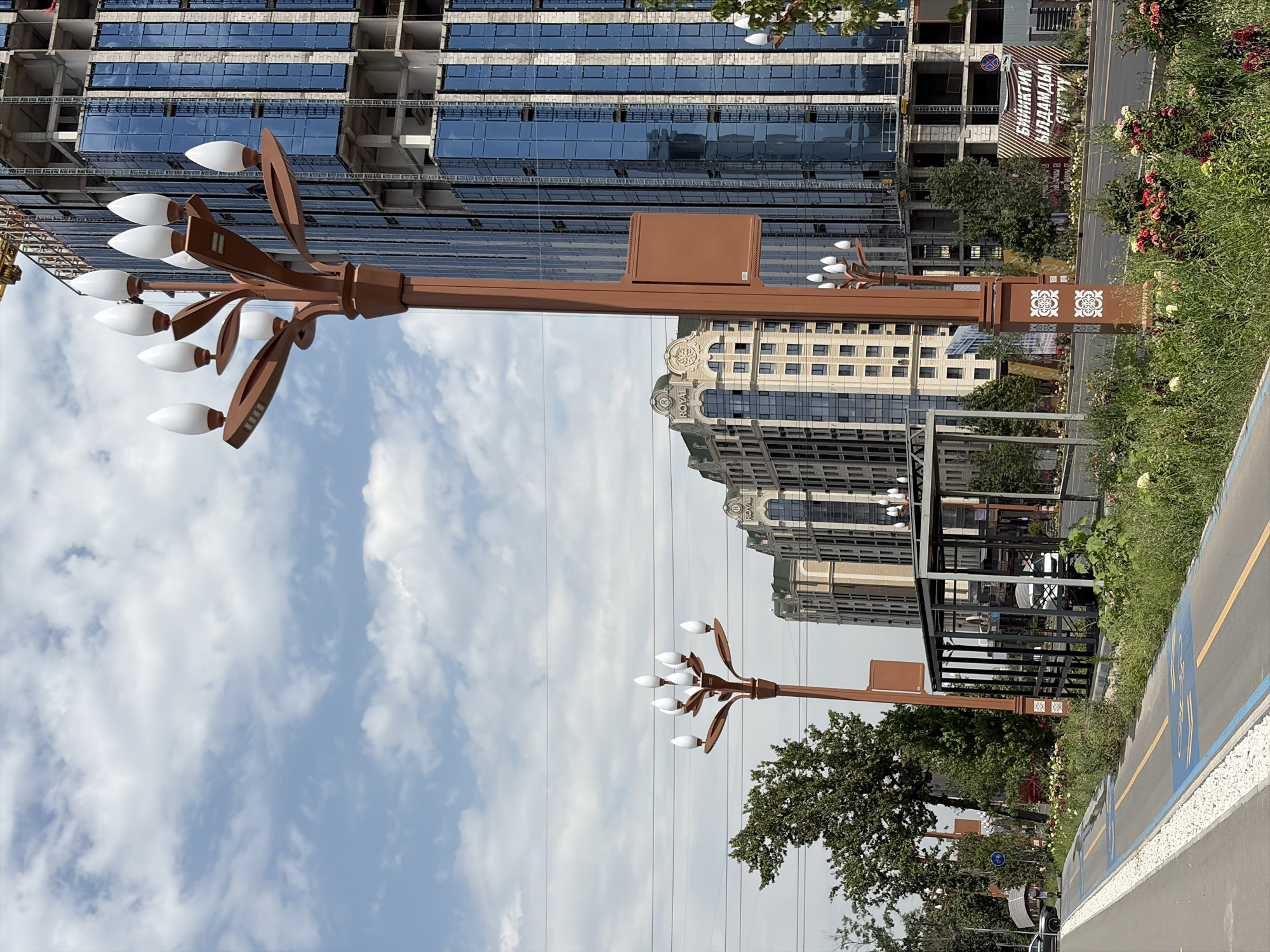 The image captures a street scene in Bishkek City, Kyrgyzstan, featuring a prominent lamppost with multiple white, tulip-shaped lights in the foreground. Behind the lamppost, a modern, multi-story building with blue glass windows is under construction, with scaffolding visible. To the left of the construction site, another, lighter-colored building with ornate detailing and the inscription "ROYAL" is visible. A dedicated bicycle lane, marked with blue and yellow lines and bicycle symbols, runs along the paved street. Greenery, including trees and flowering bushes, lines the street. A bus stop shelter is present in the midground. In the background, a cloudy sky completes the scene. A sign with text in what appears to be Cyrillic script is posted on a fence in the background.