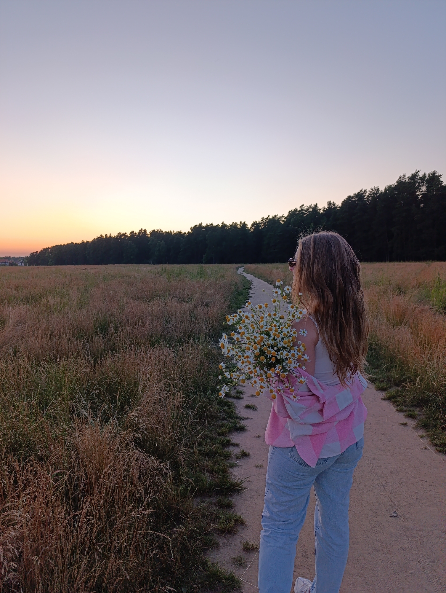 A young woman with long, blonde, wavy hair is standing on a dirt path in a grassy field at sunset. She is wearing light blue jeans and a white tank top, with a pink and white checkered jacket draped over her shoulders. She is holding a bouquet of white daisies with yellow centers. The path winds through the field and disappears into a dense line of dark green trees in the background. The sky is a gradient of pale orange and purple, transitioning to a light blue at the top. In the far distance, on the left side of the image, there are a few buildings that suggest a rural or suburban setting. The overall mood is serene and peaceful.