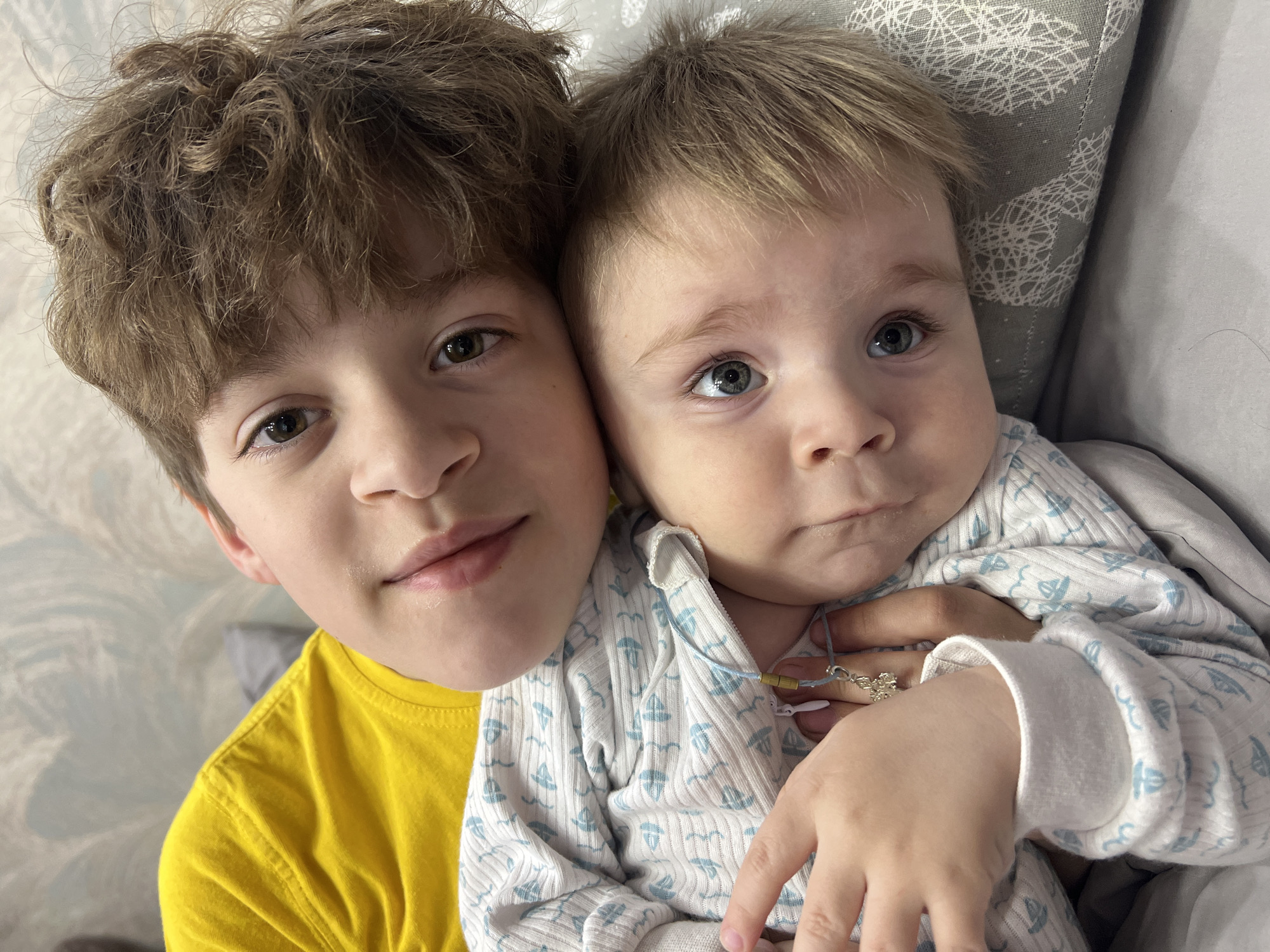 Two children are positioned close together indoors. The child on the left wears a bright yellow t-shirt and has curly brown hair. The child on the right wears a white and light blue patterned shirt. A hand is visible holding the child on the right. A pillow is in the background. The location in the city of Noviy, Russia, cannot be determined from the image.