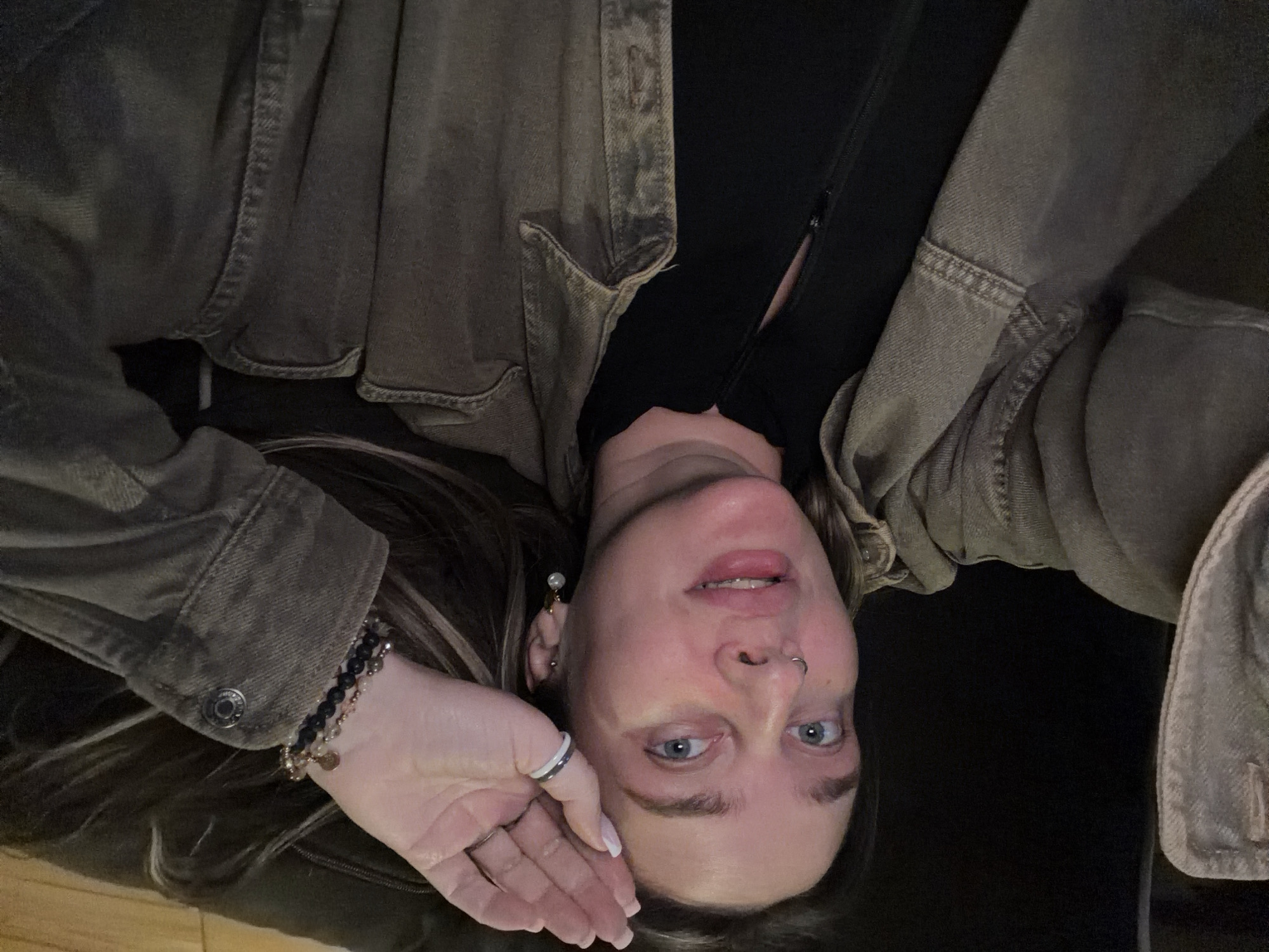 A woman with fair skin and blue eyes looks directly at the camera. She is wearing a black top underneath a brown, button-down denim jacket. She has bracelets on her left wrist and a ring on her finger. Her hair is long with blonde highlights. It is dark, and the setting is indoors. The image does not contain information to confirm its location.