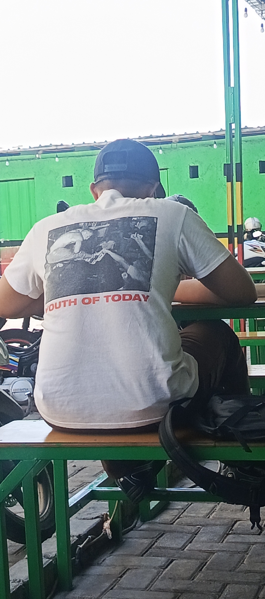 In Kletek, Indonesia, a person wearing a white t-shirt with a black and white graphic and red lettering that reads "YOUTH OF TODAY" is seated on a green metal bench. The person is also wearing a dark baseball cap. The background features a bright green wall with a yellow vertical support beam. To the right, a motorcycle rider with a white helmet is visible. Below the bench, a black backpack rests on the ground. The ground is paved with grey interlocking bricks.