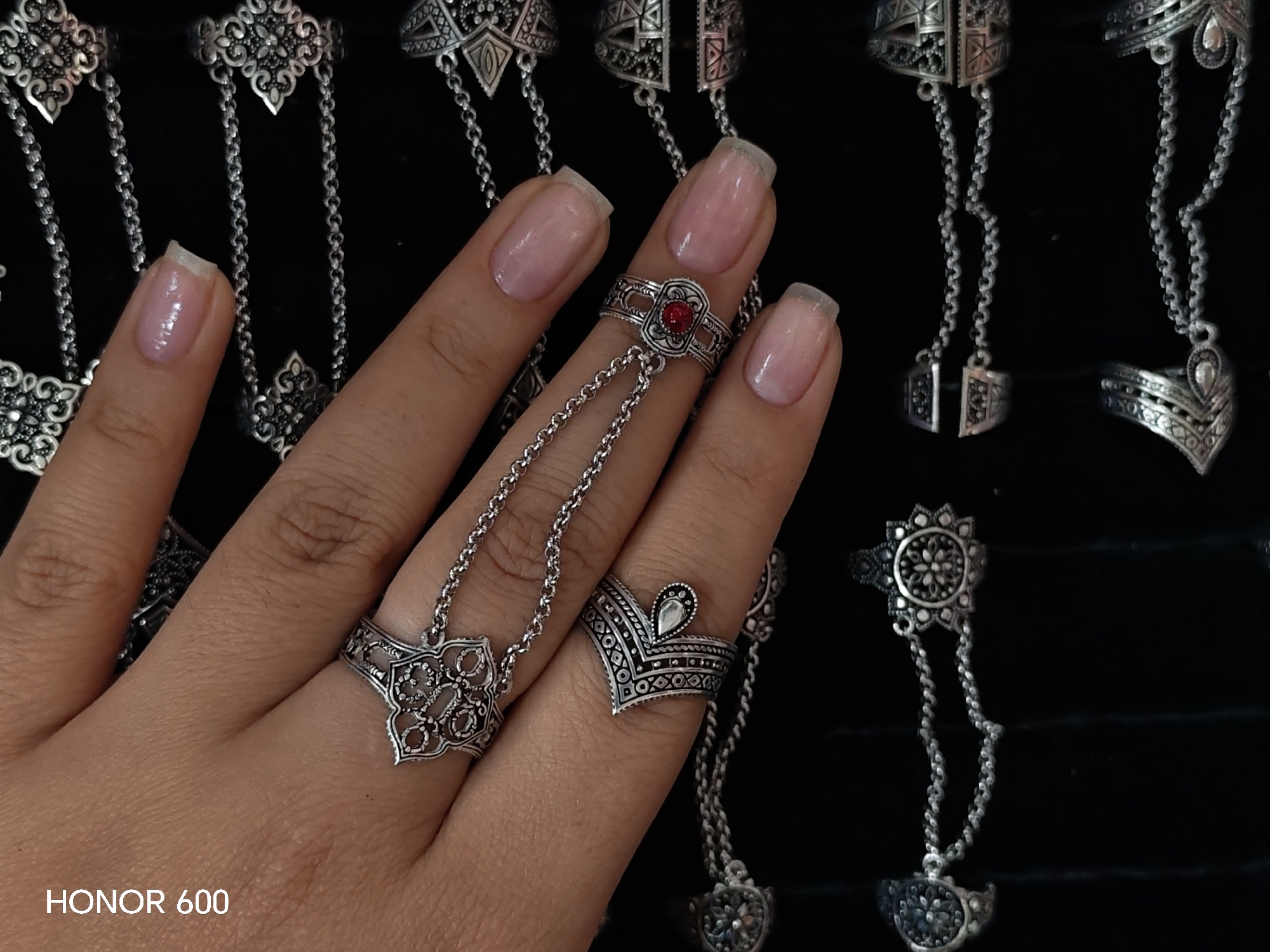 This image shows a close-up of a person's left hand, adorned with several intricate, silver-toned rings and chain jewelry. The rings are detailed with filigree, geometric patterns, and one prominent ring on the index finger features a small, red gemstone. The jewelry is designed to connect multiple rings with chains, some extending to the knuckles or beyond. The background is a dark, textured surface, possibly velvet or cloth, which serves to highlight the metallic sheen of the jewelry. In the bottom left corner, the text "HONOR 600" is visible, indicating the device used to capture the image. There is no indication of the city of Ussurijsk, Russia, or any specific location within it.