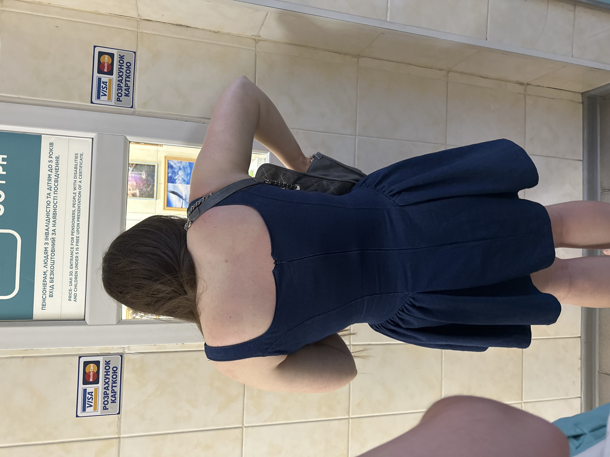 A person is standing with their back to the camera in front of a ticket vending machine. The person is wearing a dark blue dress and a grey purse is slung over their shoulder. On the wall to the left and right of the vending machine are stickers indicating that payment by Visa and Mastercard is accepted and that the machine is for ticket sales. The vending machine itself has a screen with text in Ukrainian and English. The tiled wall suggests an indoor setting, possibly a transportation hub or public facility in Odesa, Ukraine.