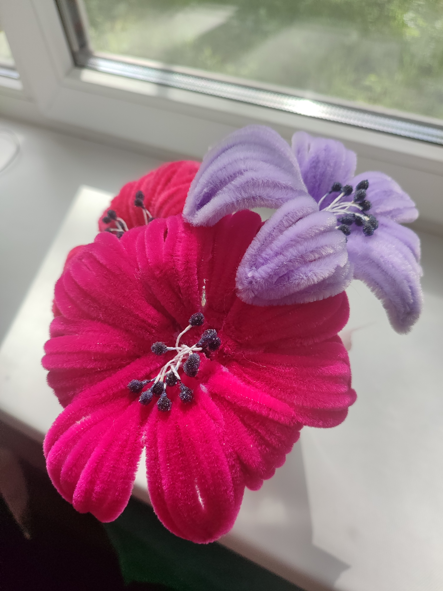 The image shows a close-up view of three artificial flowers placed on a white windowsill. The flowers are made of a fuzzy, pipe cleaner-like material. One large, bright pink flower is in the foreground, with its petals fanned out. Behind it, partially obscured, is another pink flower. To the right and slightly behind the pink flower, a lavender-colored flower with similarly fuzzy petals is visible. The center of each flower contains small, dark stamen-like structures. The background is a blurred view of a window with sunlight streaming through, suggesting an outdoor scene with greenery visible beyond the glass. The overall impression is one of a decorative arrangement.