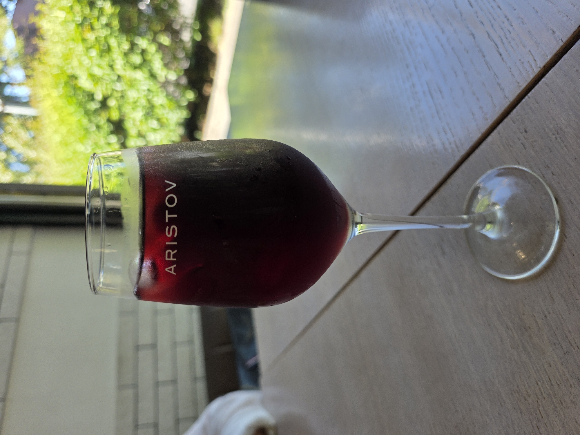 A close-up, eye-level shot showcases a glass of red liquid, likely wine or sangria, with condensation on its exterior, resting on a light-colored wooden table. The glass is tall and fluted, featuring the word "ARISTOV" etched in white on its surface. Beyond the glass, a softly blurred background reveals lush green foliage and trees, suggesting an outdoor or garden setting. The overall ambiance is serene and relaxed. This scene is set at the ARISTOV winery in the city of Sochi, Russia.