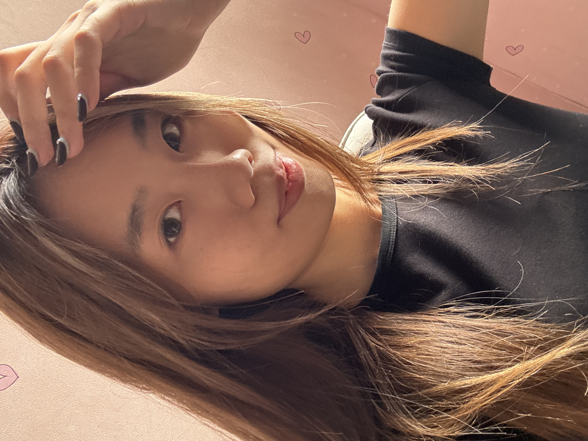 A woman with shoulder-length brown hair lies with her head tilted to the left, looking up at the camera. Her hand is raised near her head. She has dark nail polish on her fingernails. She is wearing a black top. The background is a light pink wall with small heart and lip designs. The image's location is likely indoors. The location of Shapingba District, China, cannot be confirmed from the image.
