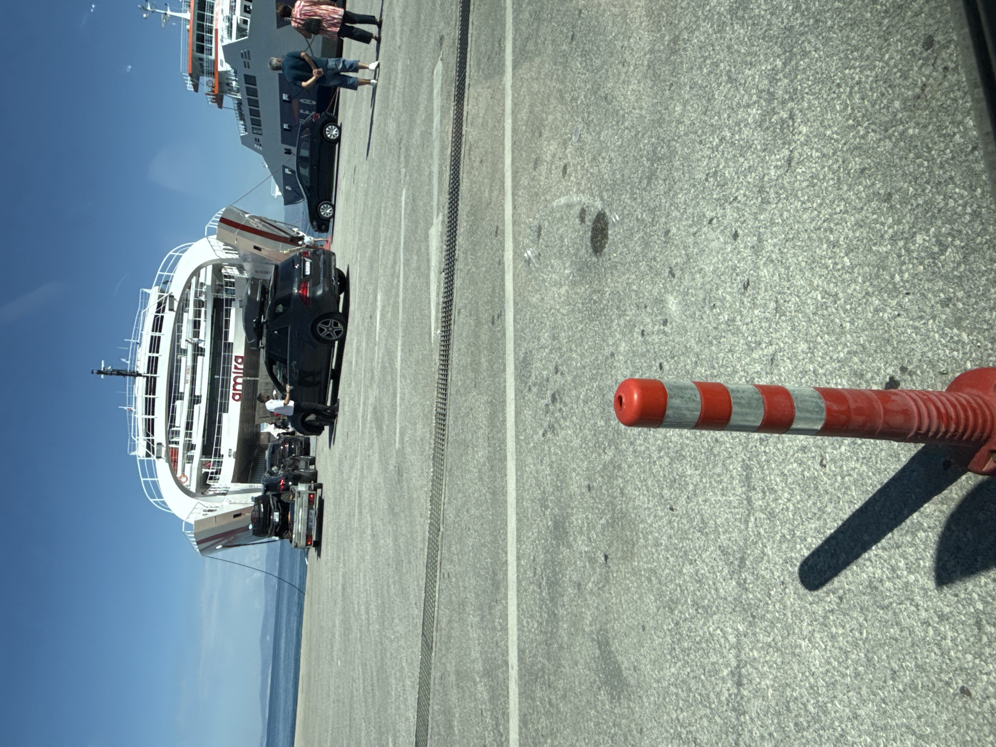 In Thassos, Greece, on a sunny day, a ferry named "AMIRA" is docked at a port, ready to embark vehicles. The ferry is large and white with red accents. In front of the ferry, several cars are lined up on the concrete pier, waiting to board. One car is on a trailer, carrying what appears to be a motorcycle or ATV. A man in a white shirt and dark pants stands near one of the cars, possibly a port worker. Further to the right, another ferry is docked, its upper decks visible. Two people, a man in a blue shirt and shorts and a woman in a patterned top and dark pants, stand observing the scene. In the foreground, a red and white striped traffic cone is positioned on the ground, casting a long shadow. The sky is a clear blue, and the sea is calm.